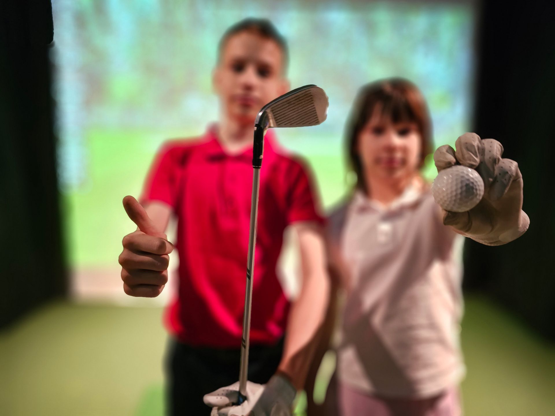 A boy and girl hold a golf club and golf ball, respectively, at a golf simulator. A boy and girl hold a golf club and golf ball, respectively, at a golf simulator.