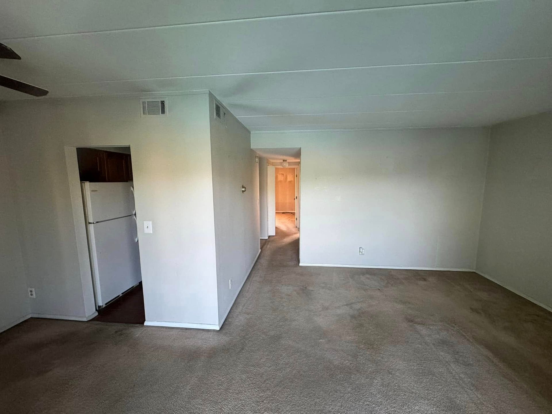 An empty living room with a refrigerator and a ceiling fan.