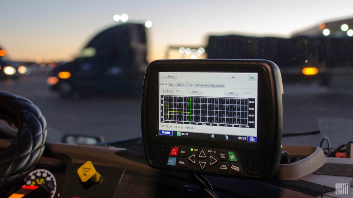 An electronic logging device (ELD) in a truck cab displays data, with trucks and a sunset in the blurred background.
