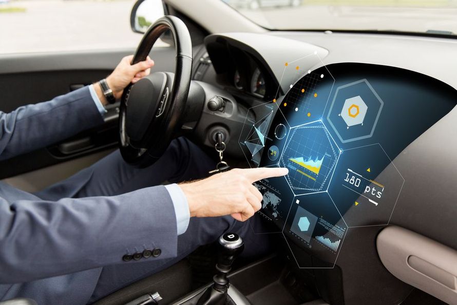 A man in a suit is driving a car and pointing at the dashboard.