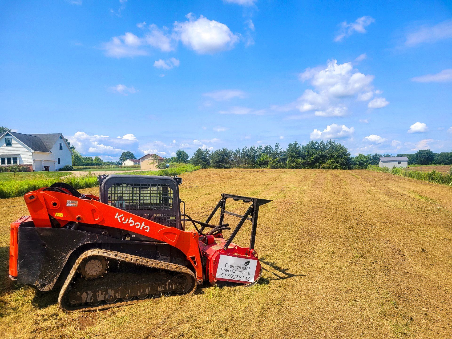 Red Kubota skid steer with a brush cutter mulching a field on a sunny day.