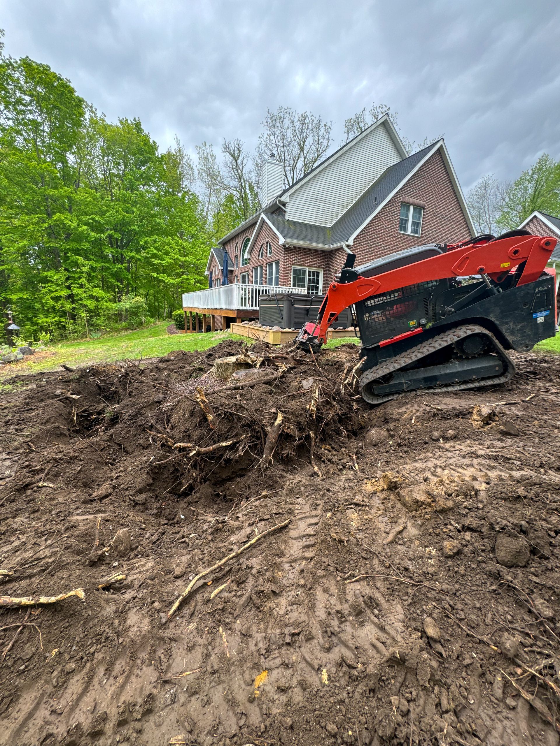 A small red and black skid steer removing tree stumps in a backyard with a brick house.