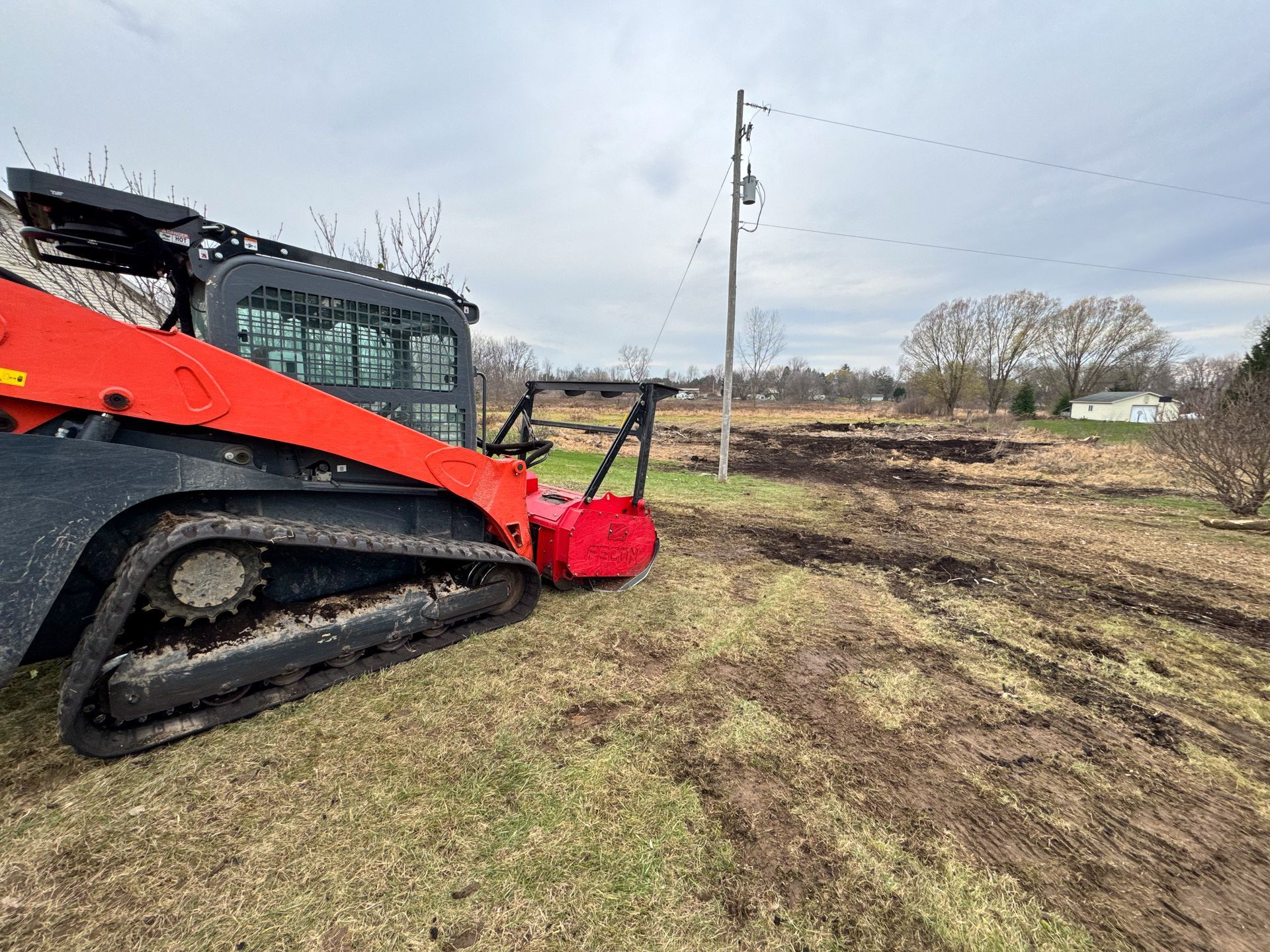 Orange track loader with brush cutter clearing land; overcast day.
