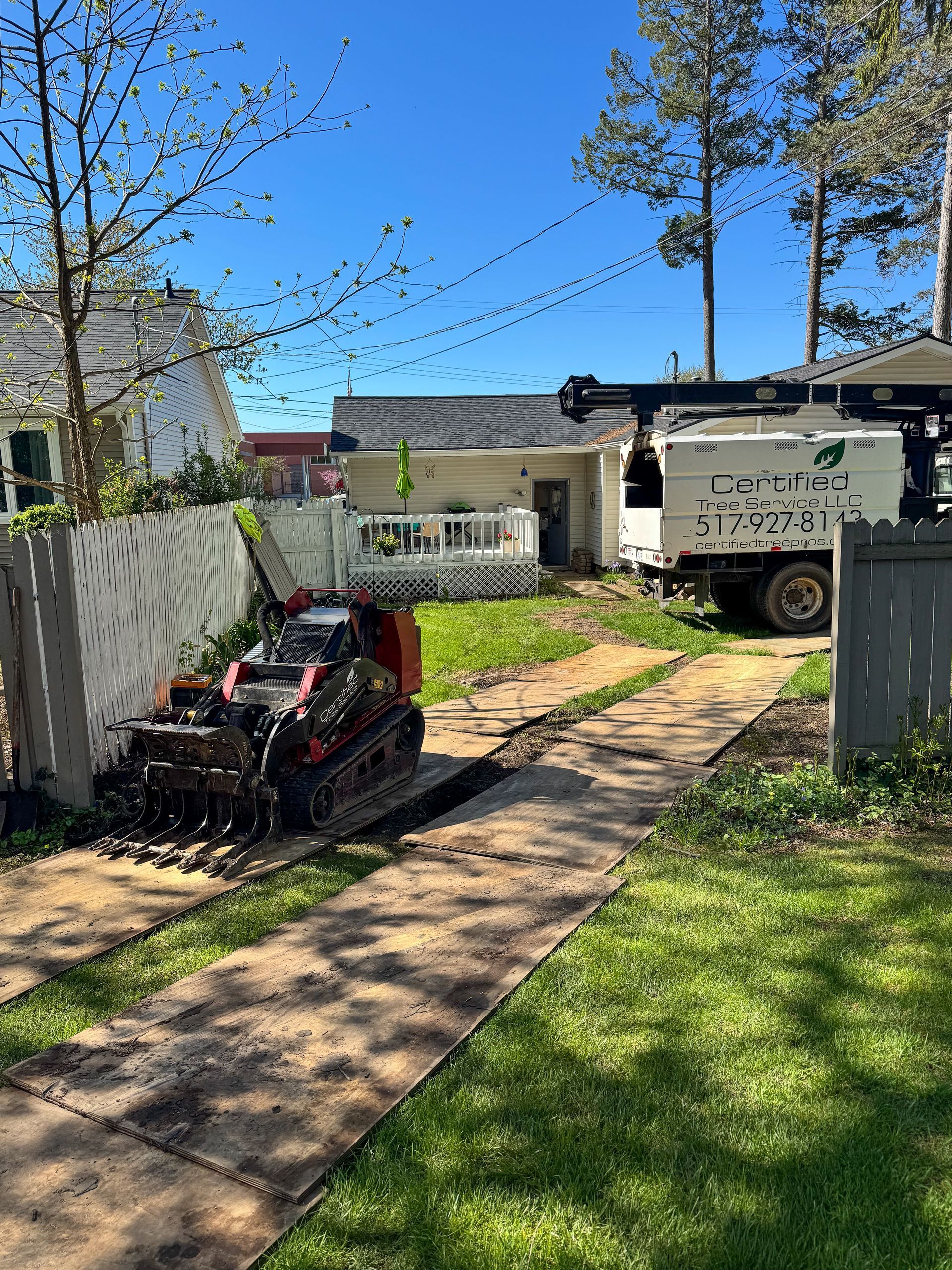 A mini excavator grinds asphalt on a driveway. Behind it is a house and a tree service truck.