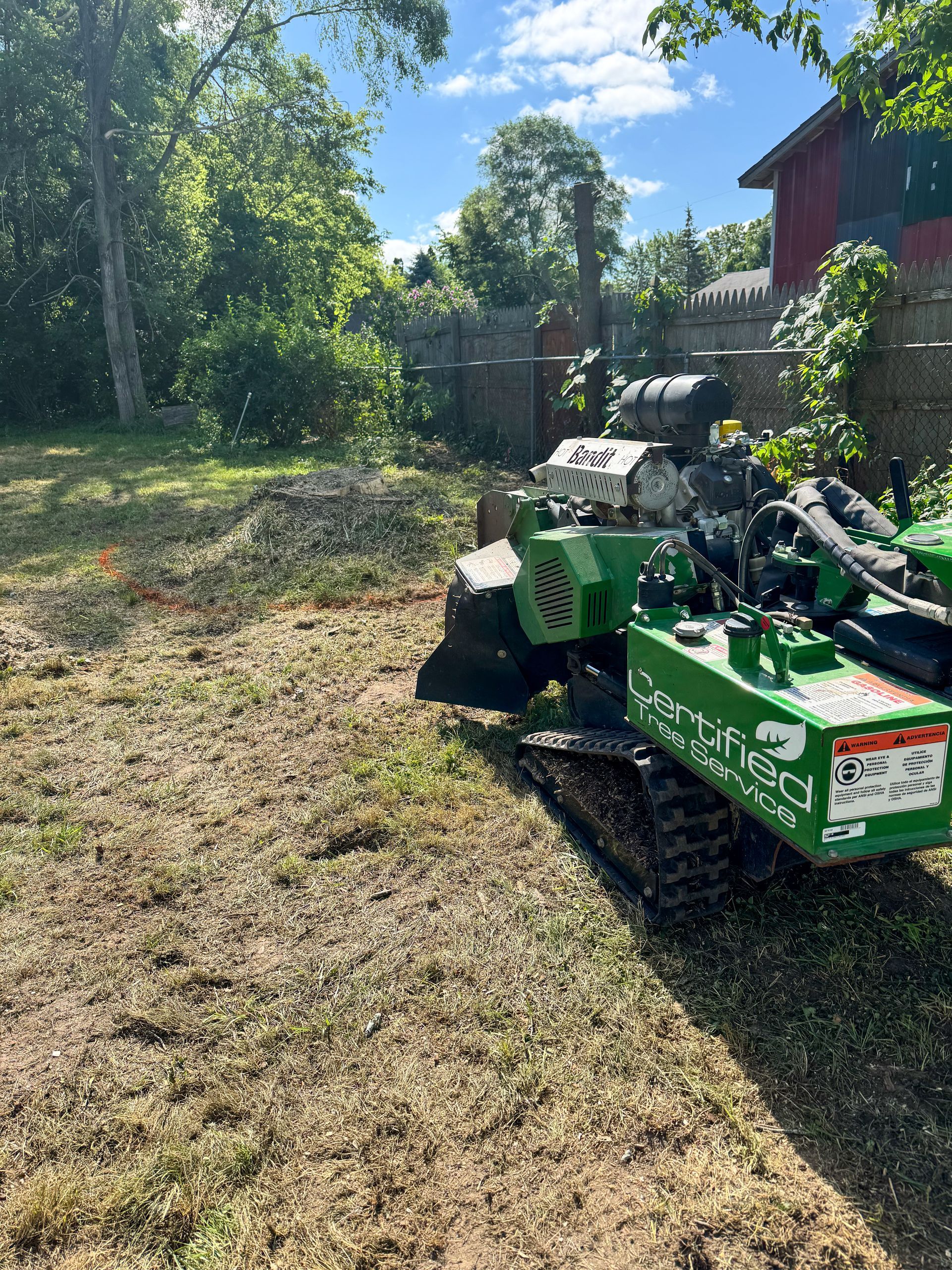 A green stump grinder on a lawn, trees, and a red building in the background on a sunny day.