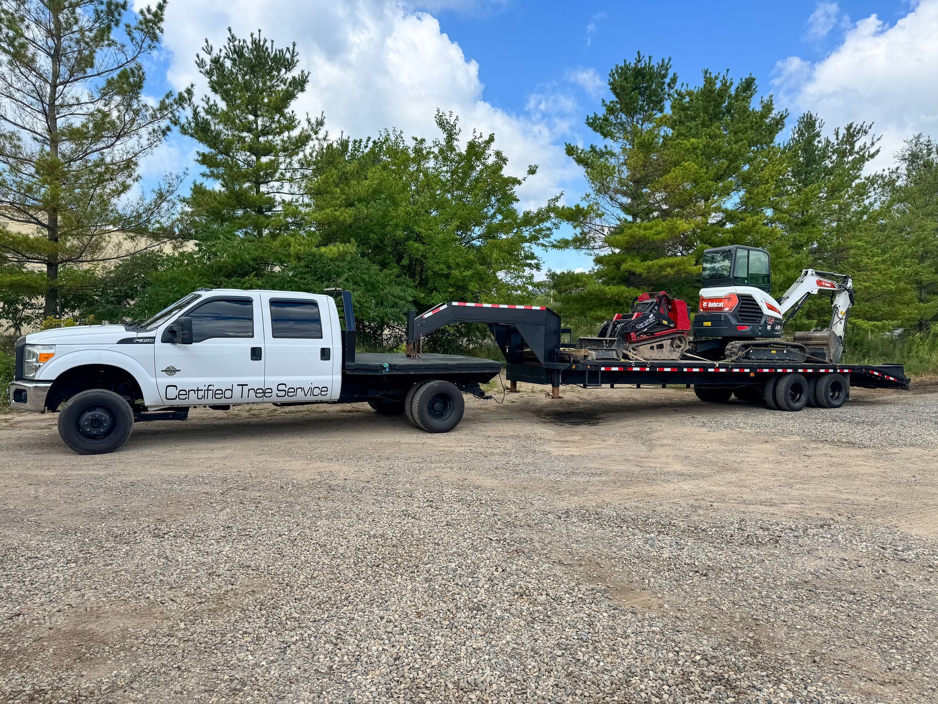 White truck pulling a trailer with an excavator on it. Blue sky. Gravel lot.