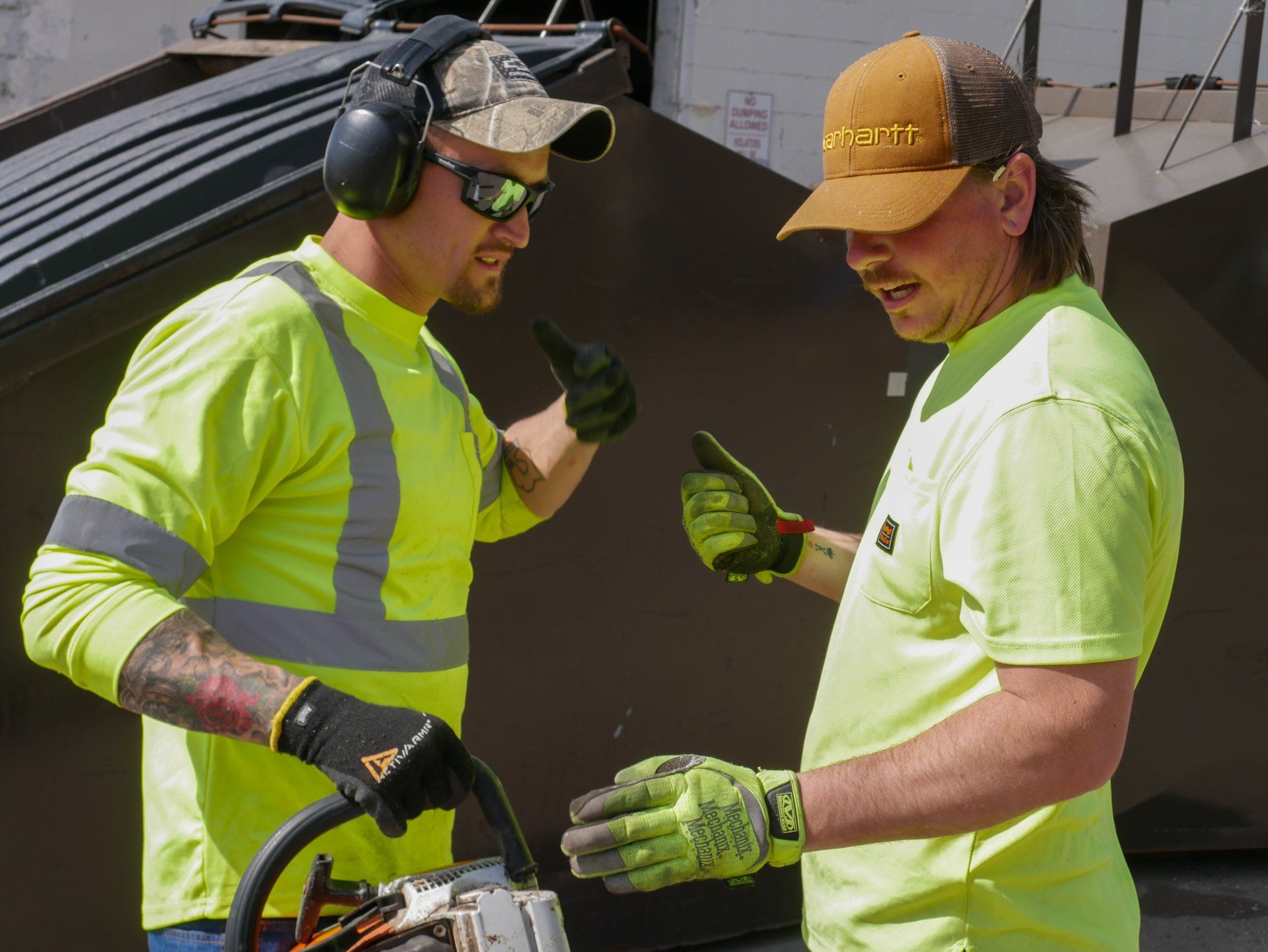 Two construction workers are talking to each other while one is using a chainsaw.