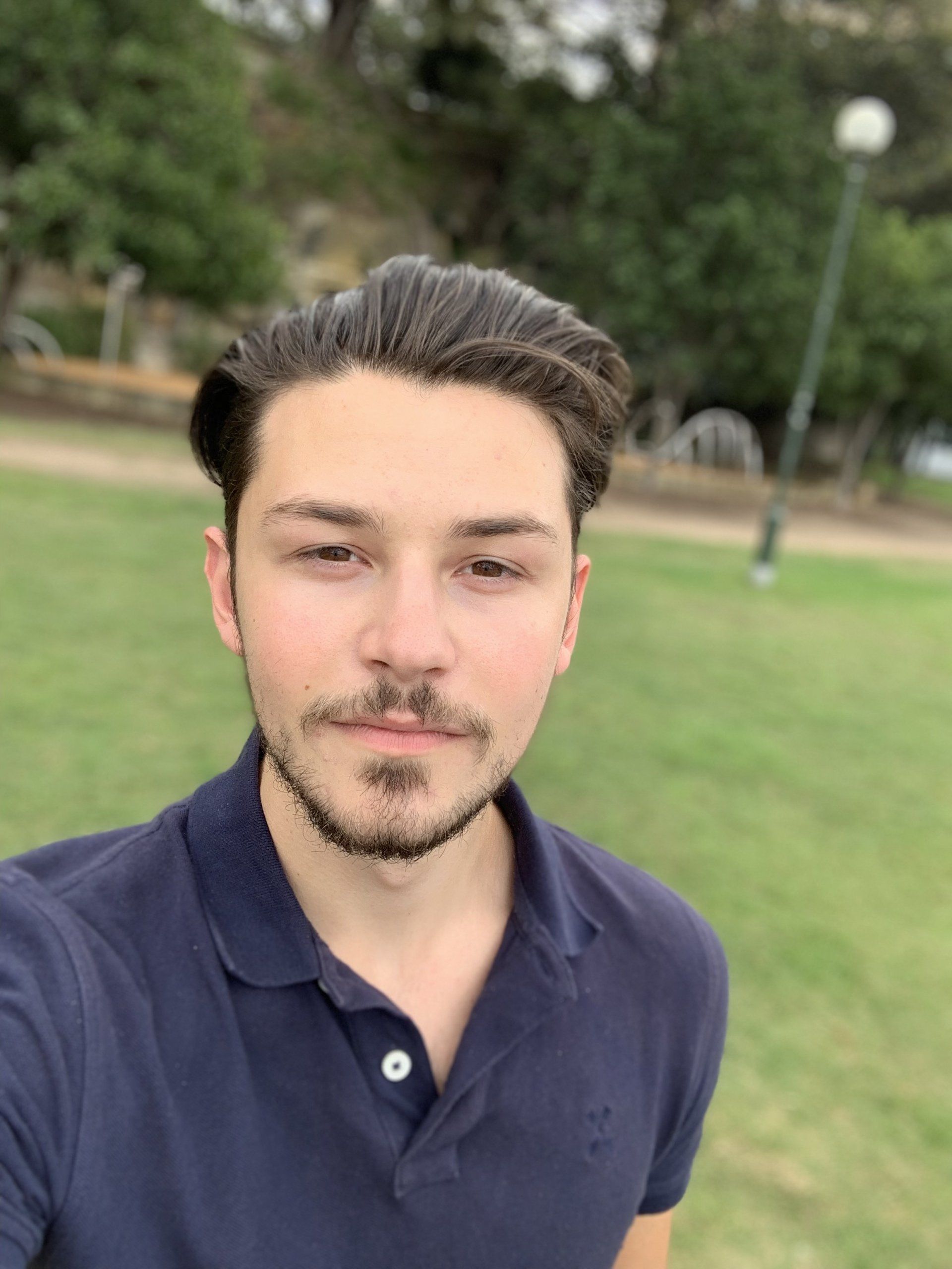 A young man with a beard is taking a selfie in a park.