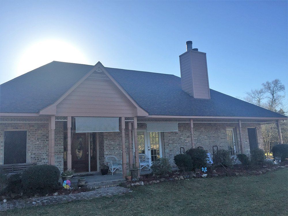 Brick house with dark roof and chimney, porch with rolled-up shades, and sunny sky.