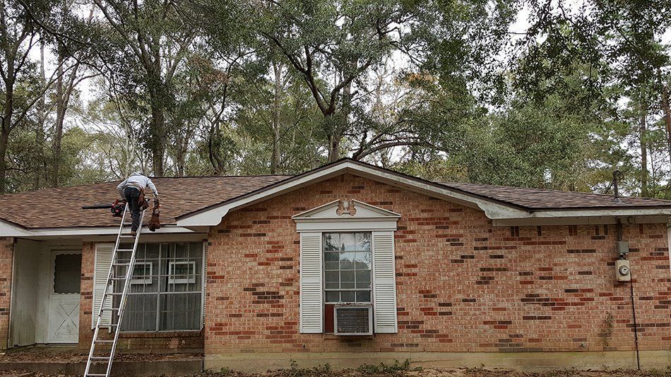 Man on ladder repairing roof of brick house surrounded by trees.
