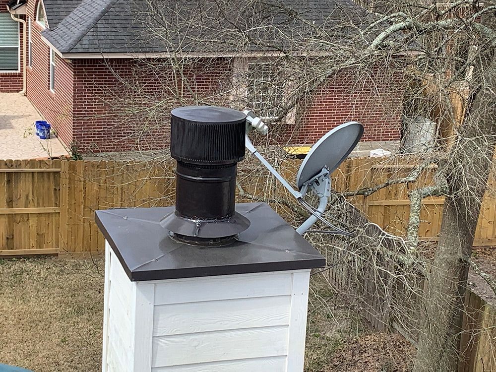 Chimney with black cap and satellite dish on a house roof. White siding, brown brick background.