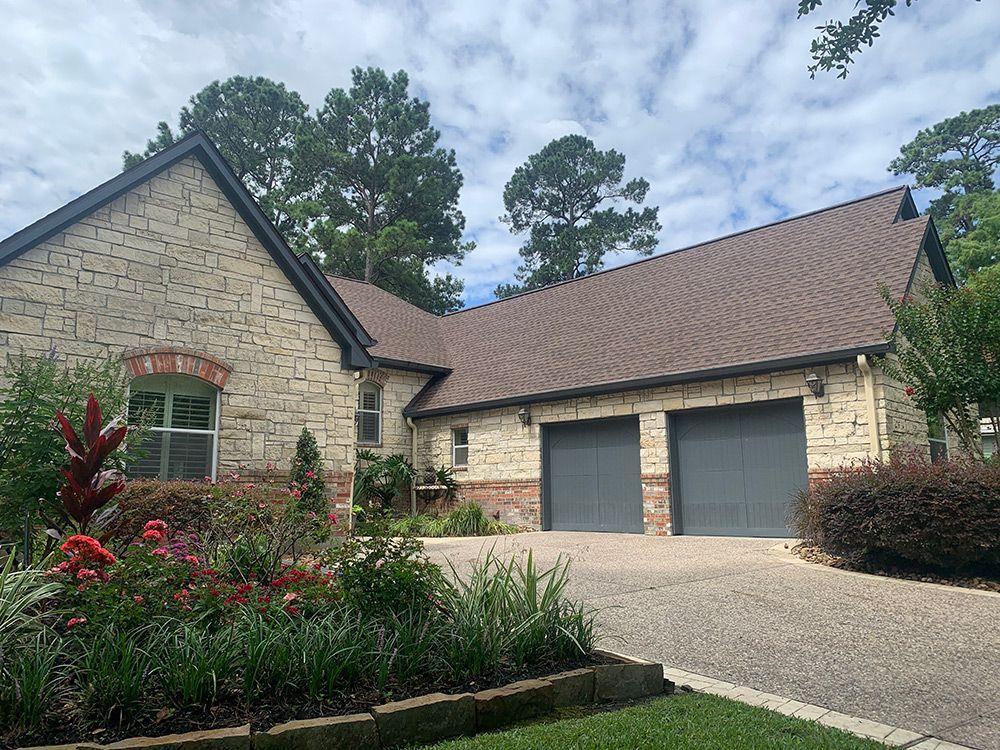 Tan brick house with a brown roof and gray garage doors. Garden in front, driveway leading up.