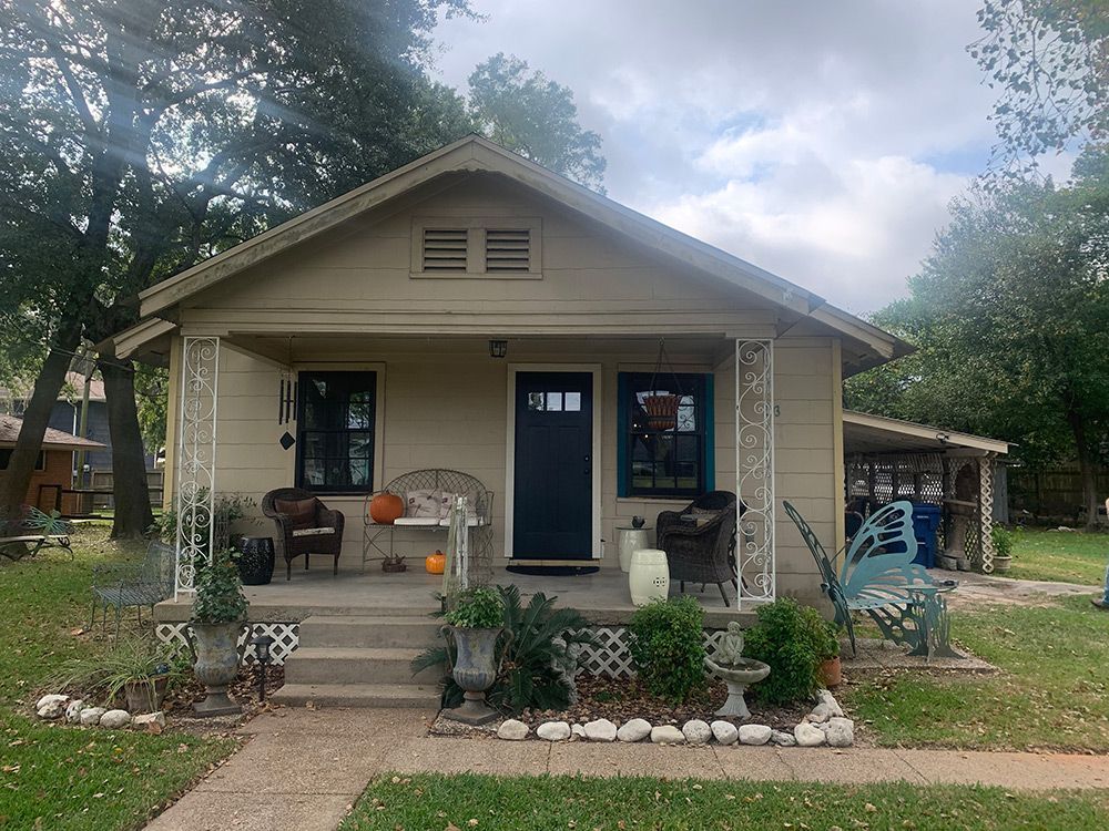 Tan cottage with dark blue door, small porch with stone steps, flanked by flower beds and seating.