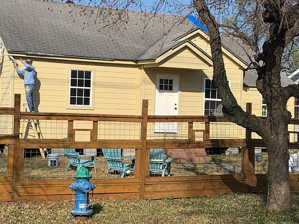 Person painting a yellow house from a ladder, with a wooden fence and blue chairs in front.