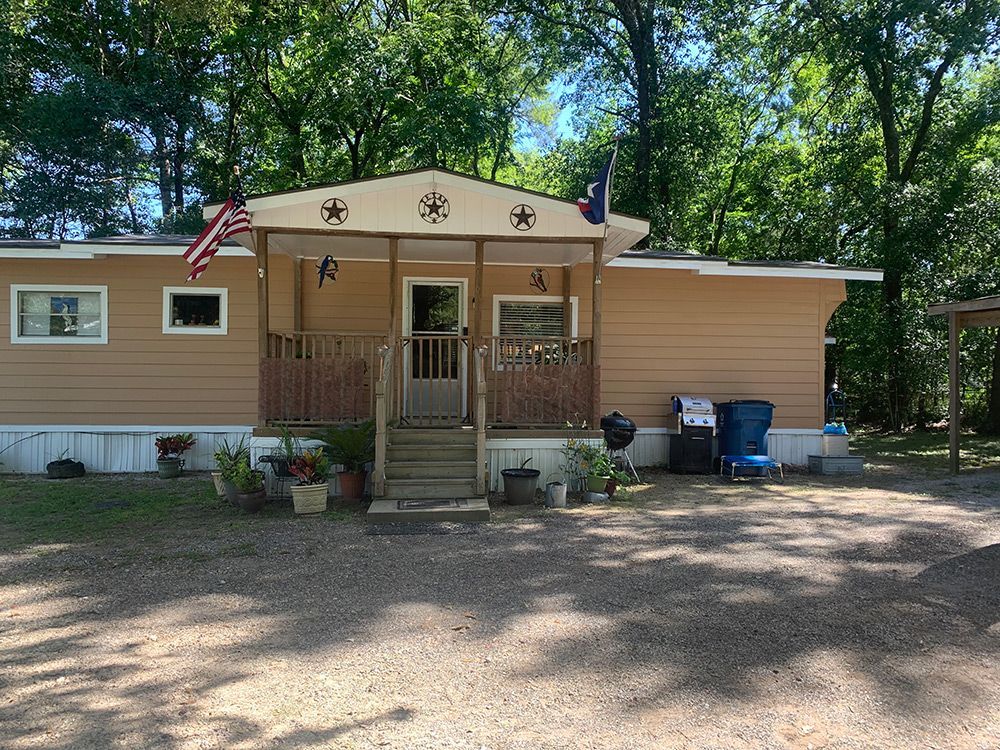 Tan mobile home with small porch, American flags, and a gravel driveway, in a wooded area.