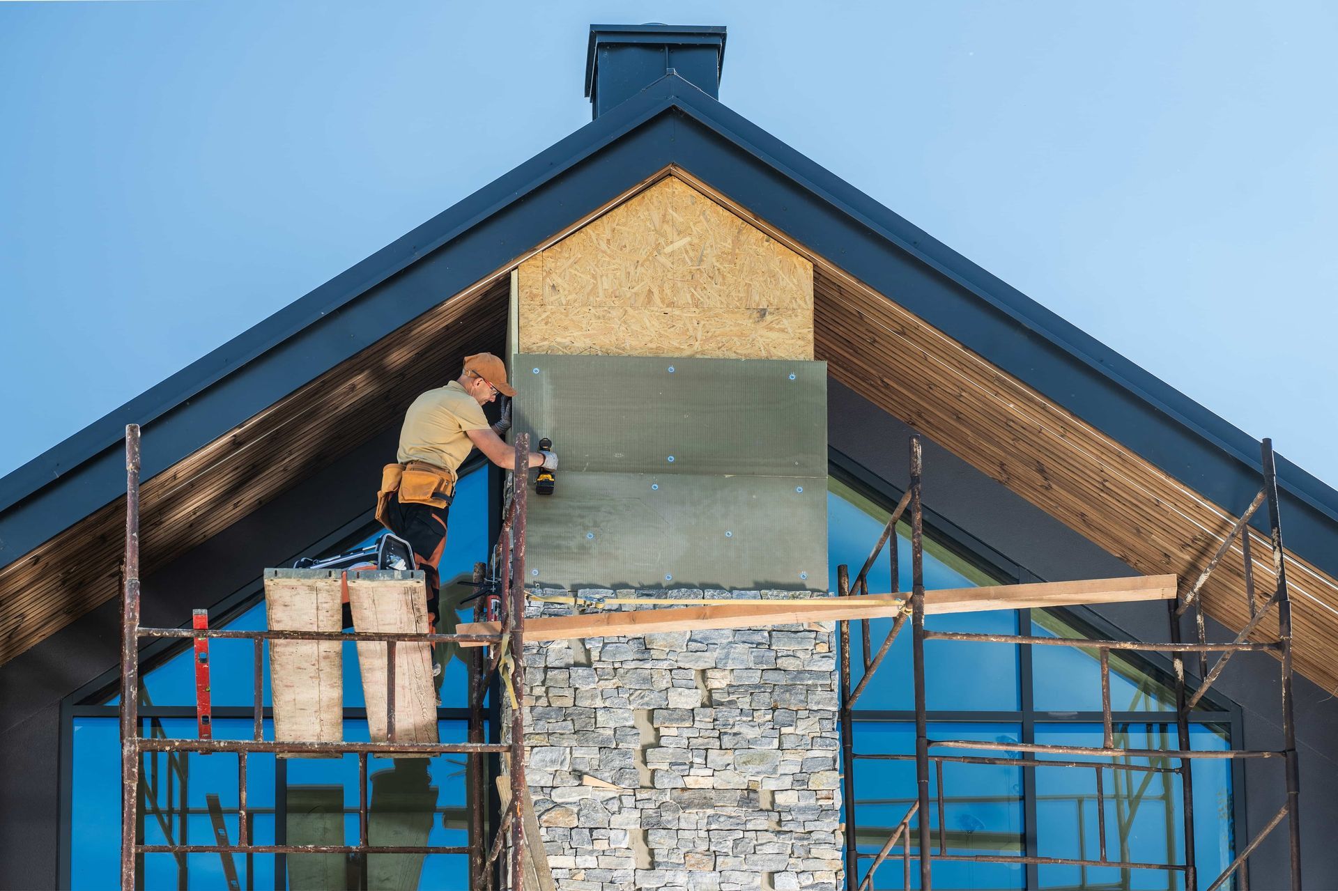 Construction worker on scaffolding installing siding on a chimney. Bright blue sky.