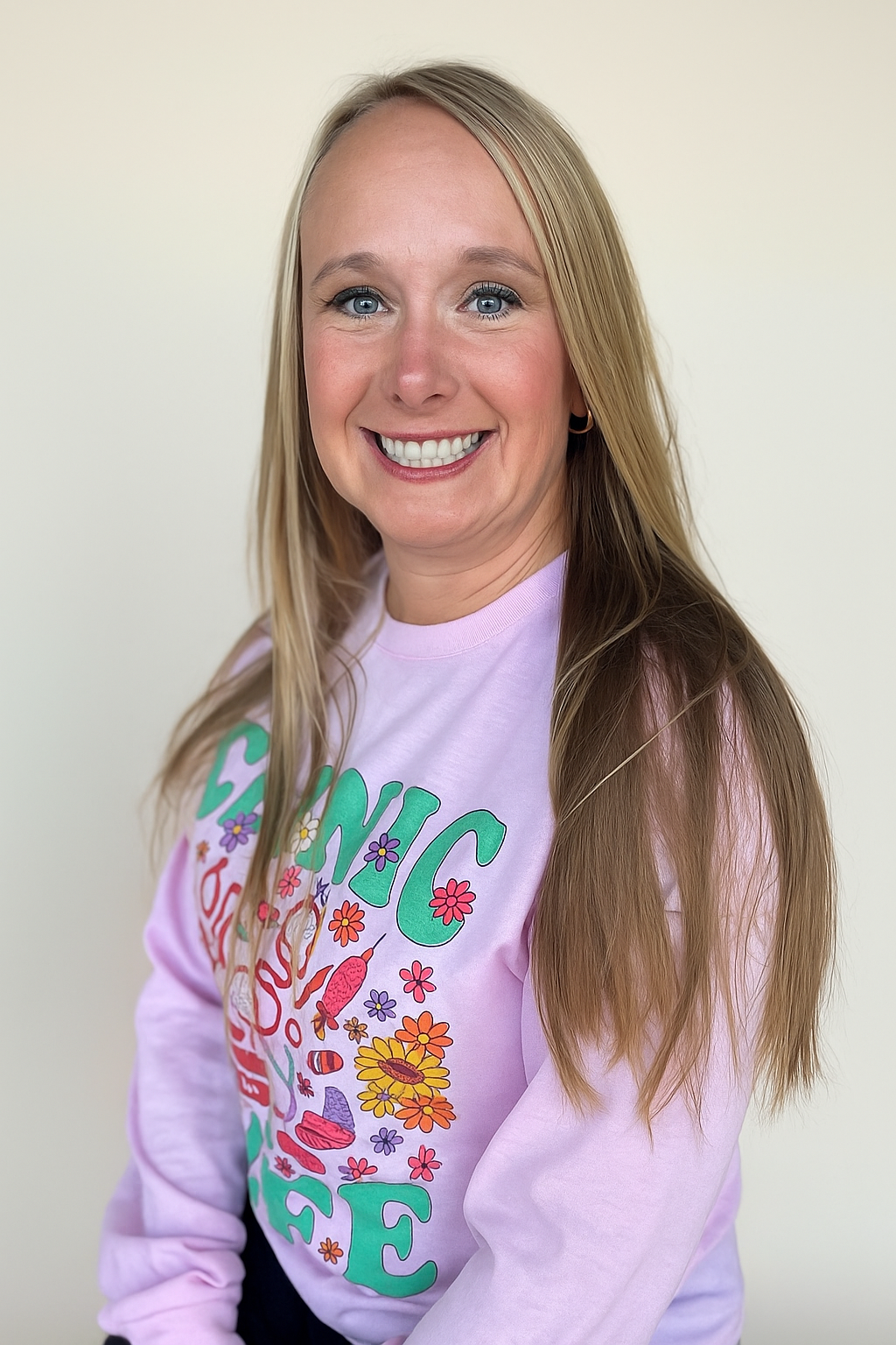 Woman in a pink sweater smiles, blonde hair, light background. Sweater says 