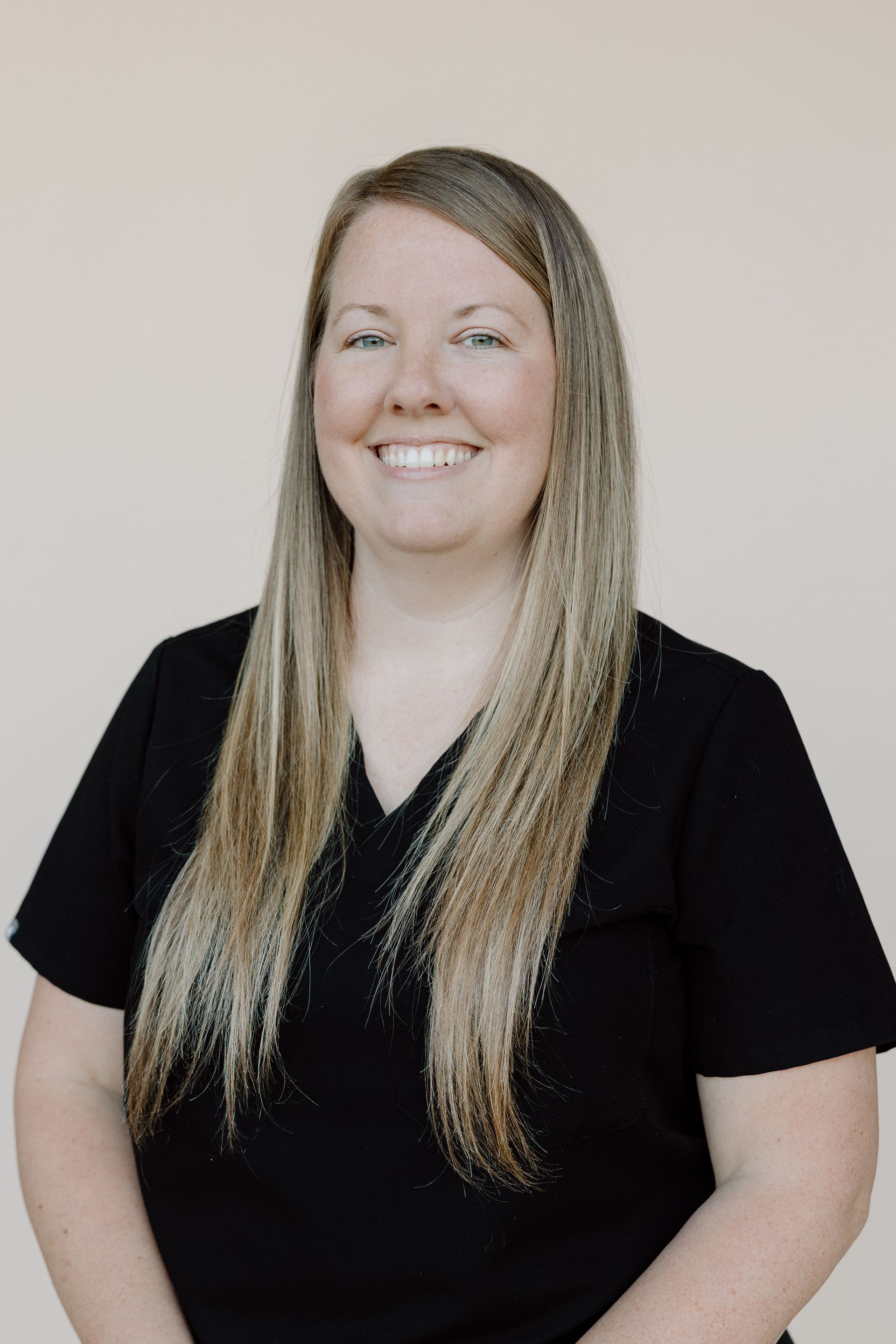 Woman with long blonde hair, smiling, wearing a black top, against a beige background.