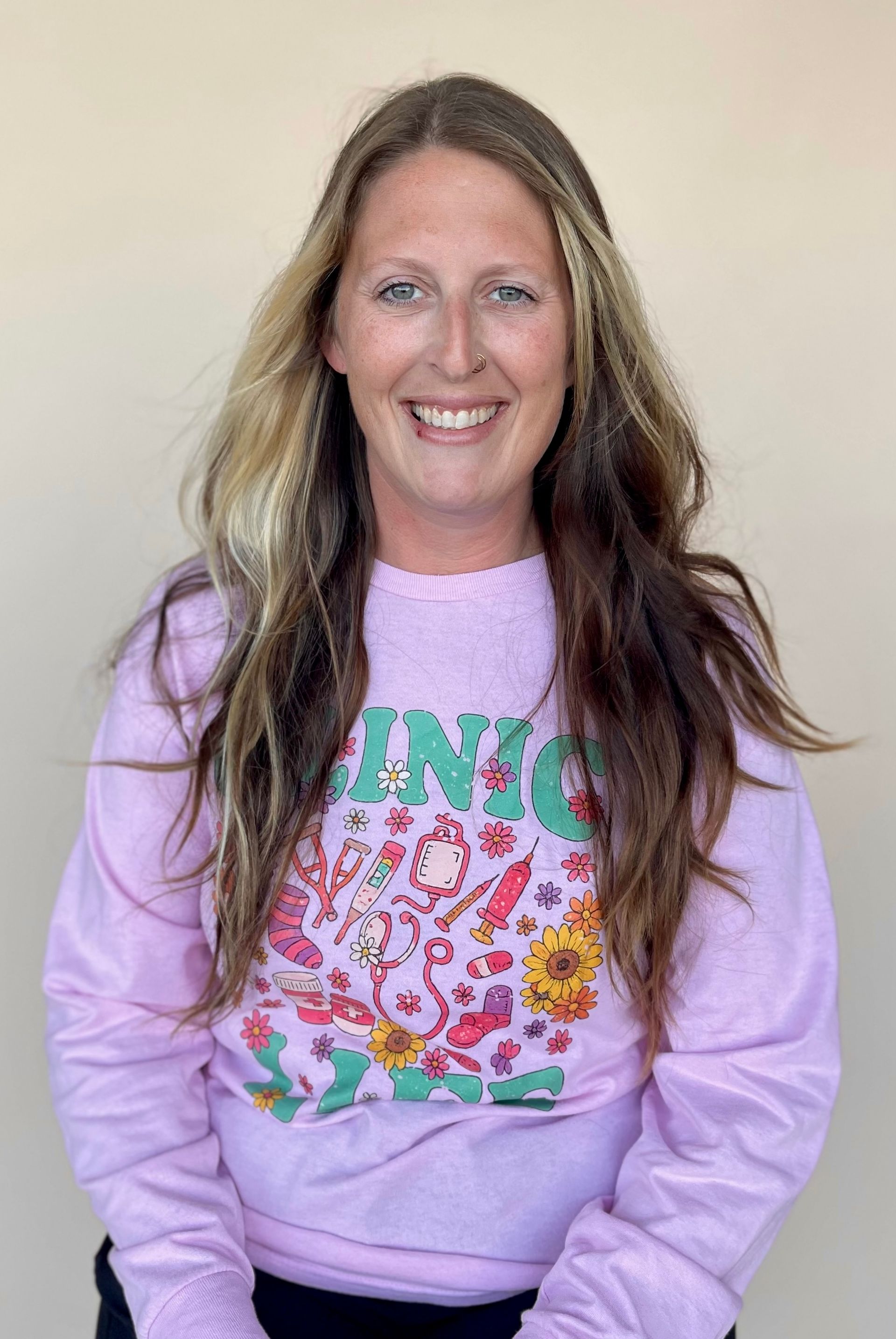 Woman with long, wavy hair, smiling, wearing a pink sweatshirt with a colorful graphic.