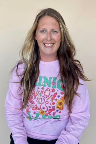 Woman with long, wavy hair, smiling, wearing a pink sweatshirt with a colorful graphic.