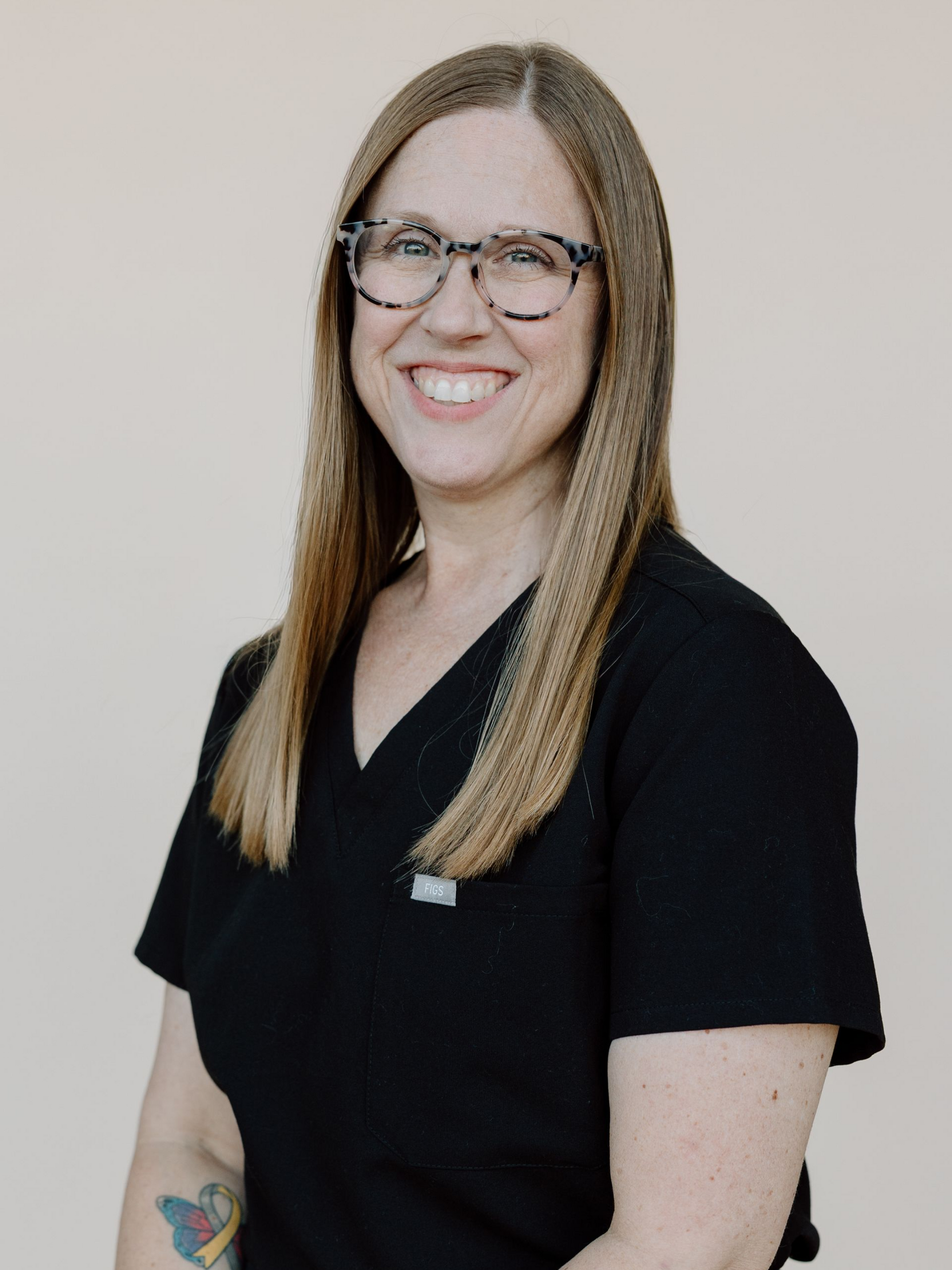 Woman with glasses smiles, wearing black scrubs, standing against a beige background.