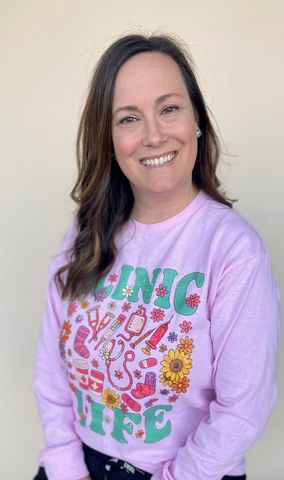Woman smiling, wearing a pink “Clinic Life” sweatshirt, standing in front of a neutral-colored background.