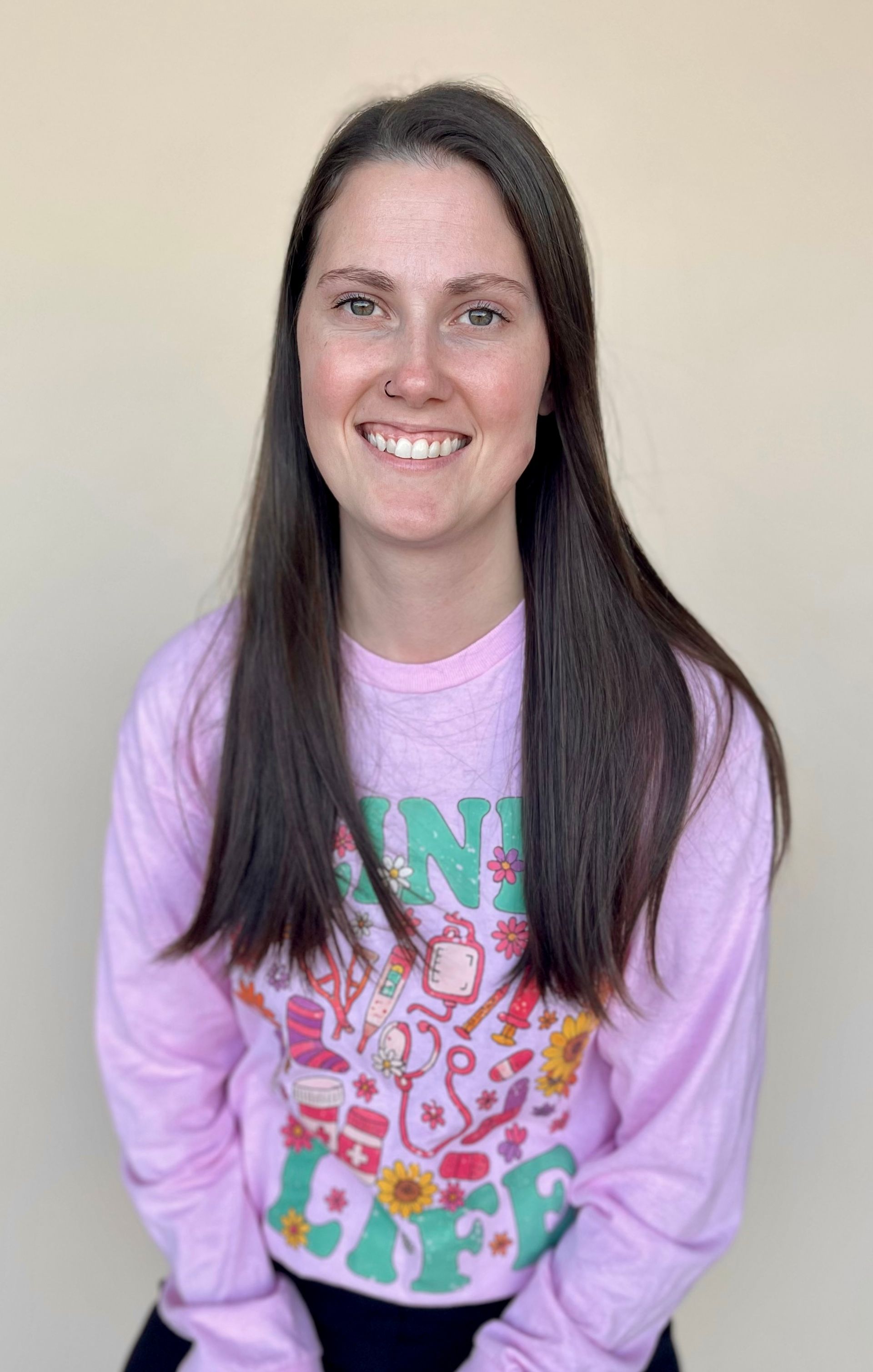 Woman smiling, wearing a pink long-sleeved shirt with colorful graphic and standing against a tan wall.