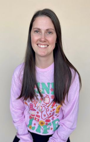 Woman smiling, wearing a pink long-sleeved shirt with colorful graphic and standing against a tan wall.