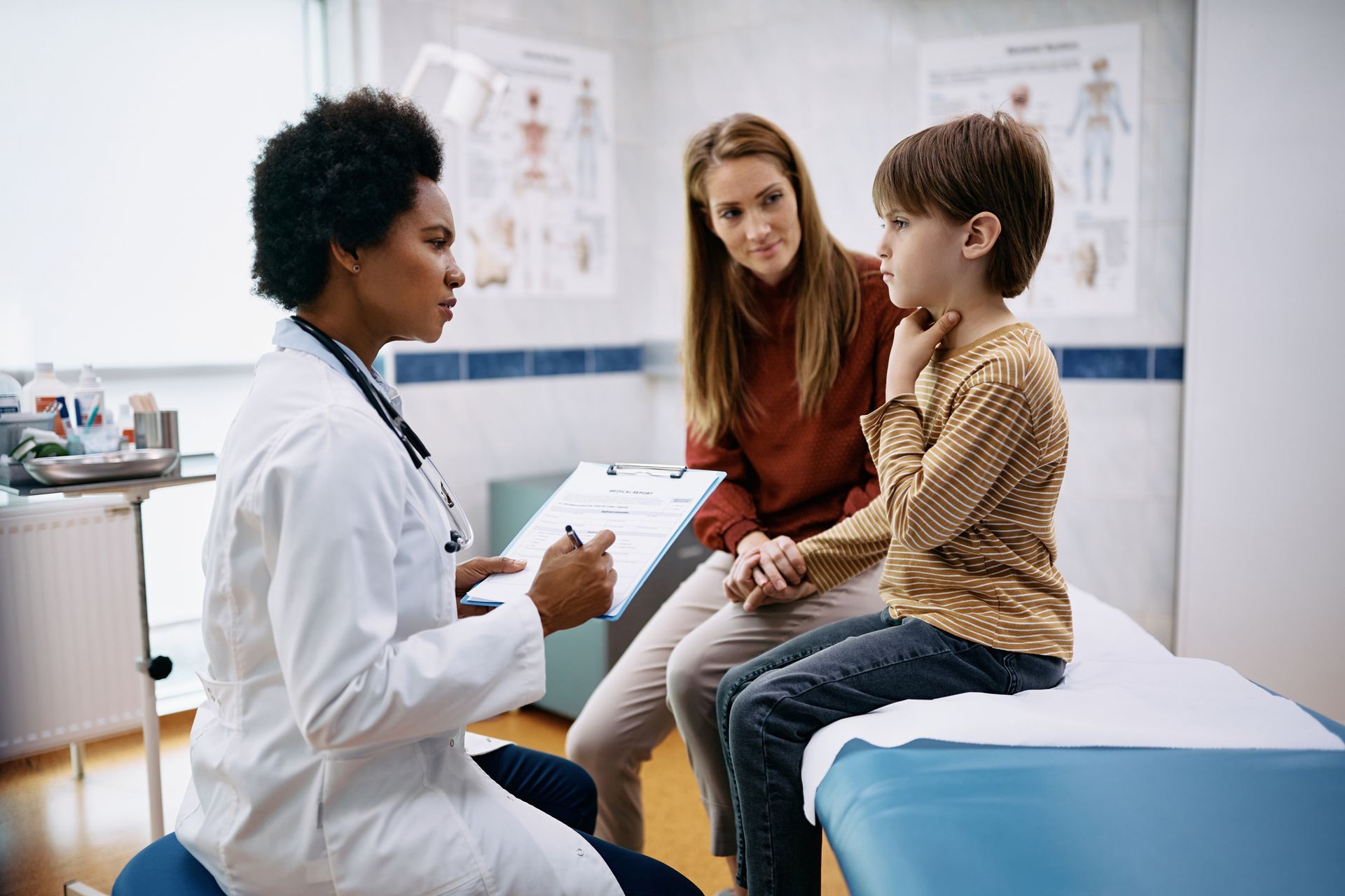 Doctor examines a child's throat, mother present. Doctor wears white coat, boy sits on a bed, and the mom watches.