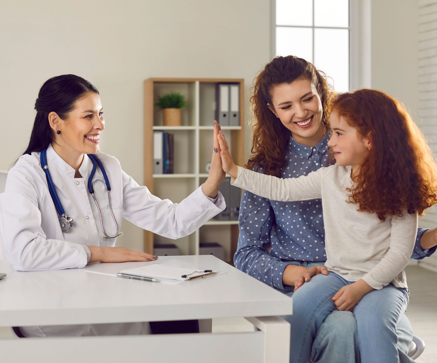Doctor high-fives a child sitting with their parent in a medical office, all smiling.