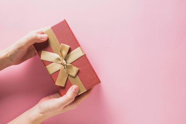 A woman is holding a red gift box with a gold ribbon on a pink background.