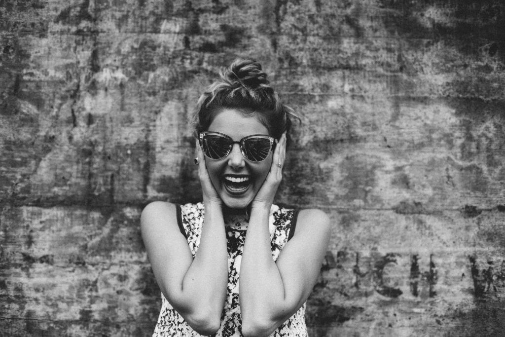 A woman wearing sunglasses is covering her ears in front of a brick wall.