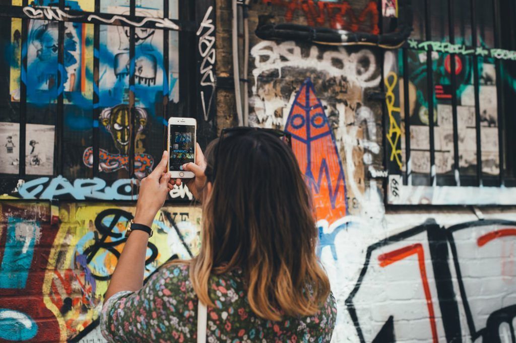 A woman is taking a picture of a graffiti covered wall with her cell phone.