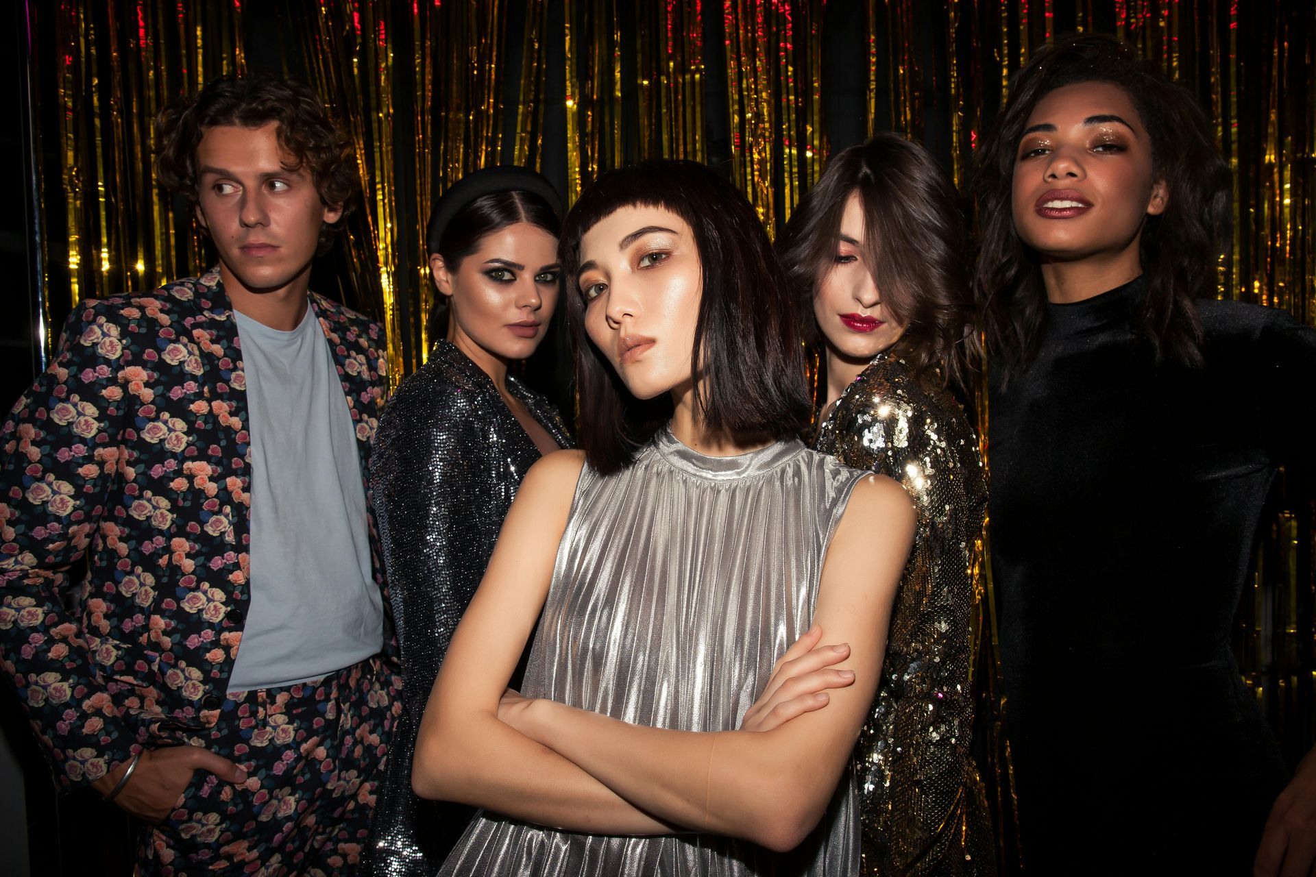 Group of five people posing in front of gold tinsel. Woman in center with silver dress and arms crossed. Others in sparkly outfits.
