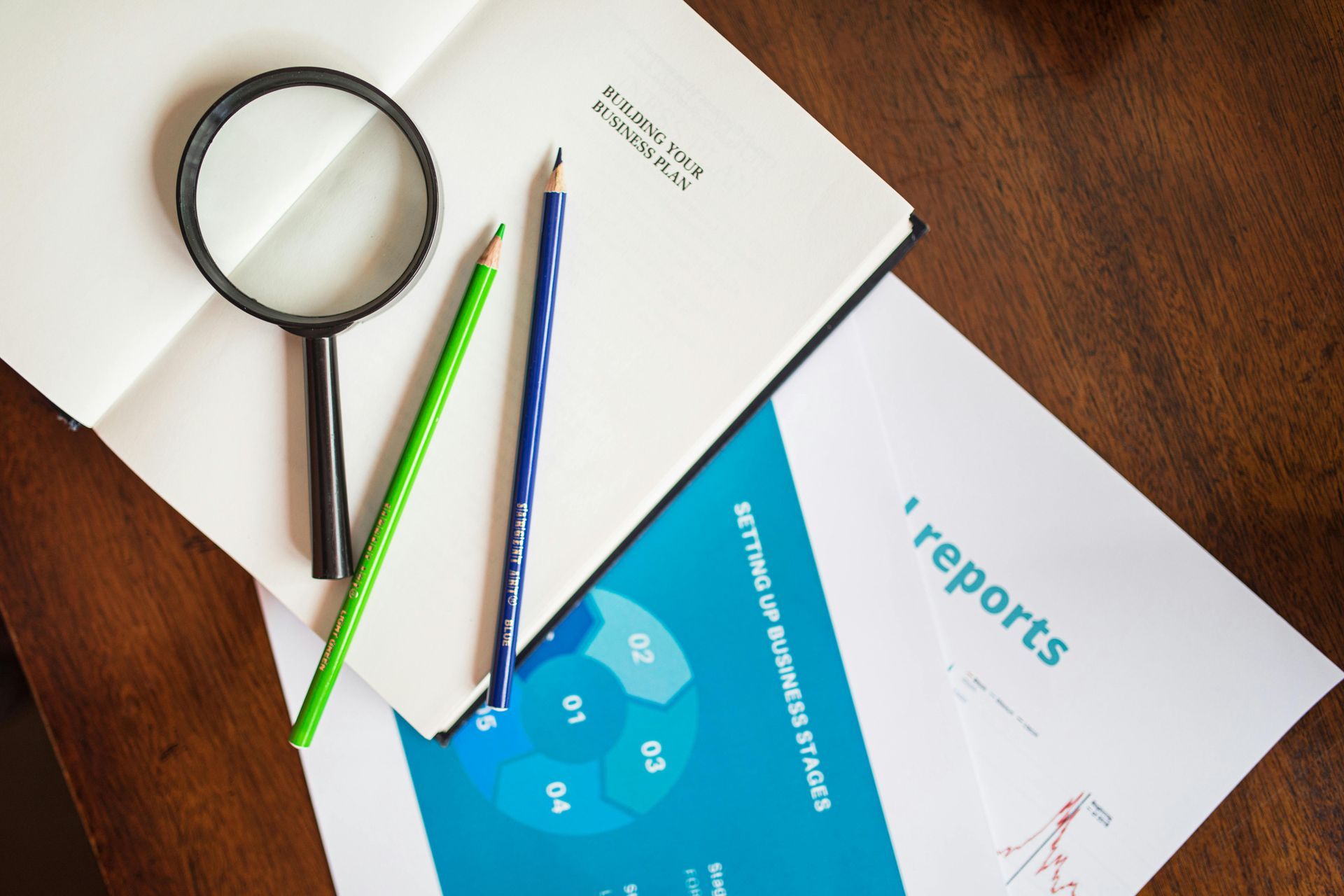 Lupa, lápis e documentos sobre uma mesa de madeira; plano de negócios e relatórios para análise.