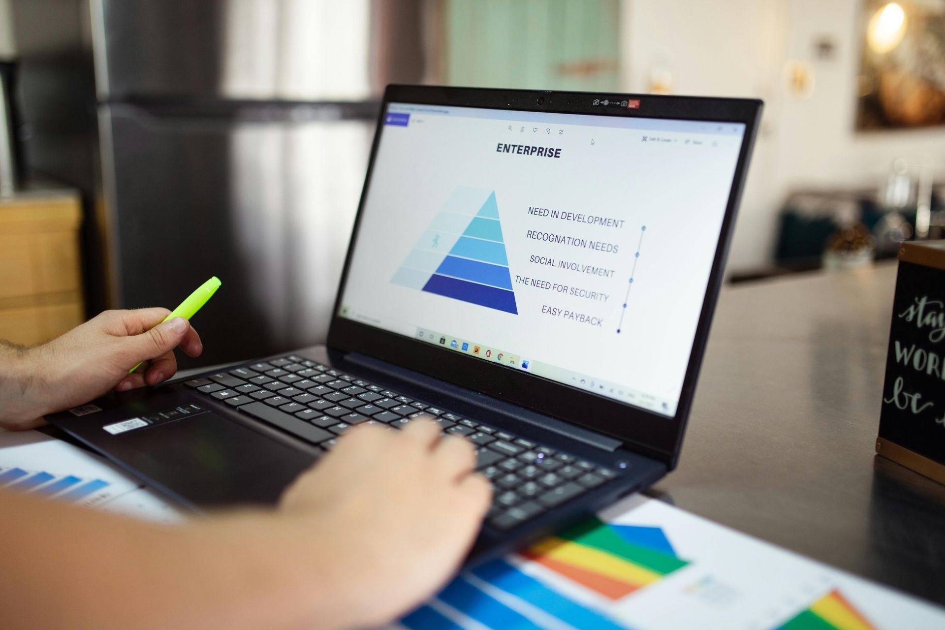 Person working on a laptop, viewing a pyramid chart; a green highlighter and charts are nearby.