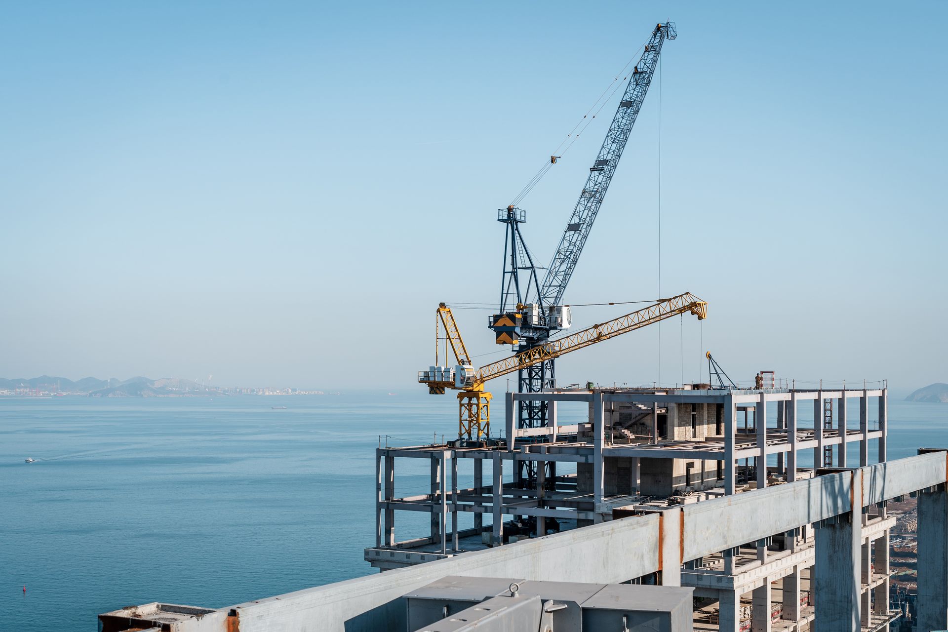 Construction site with two cranes over a building frame against a blue sea and sky.