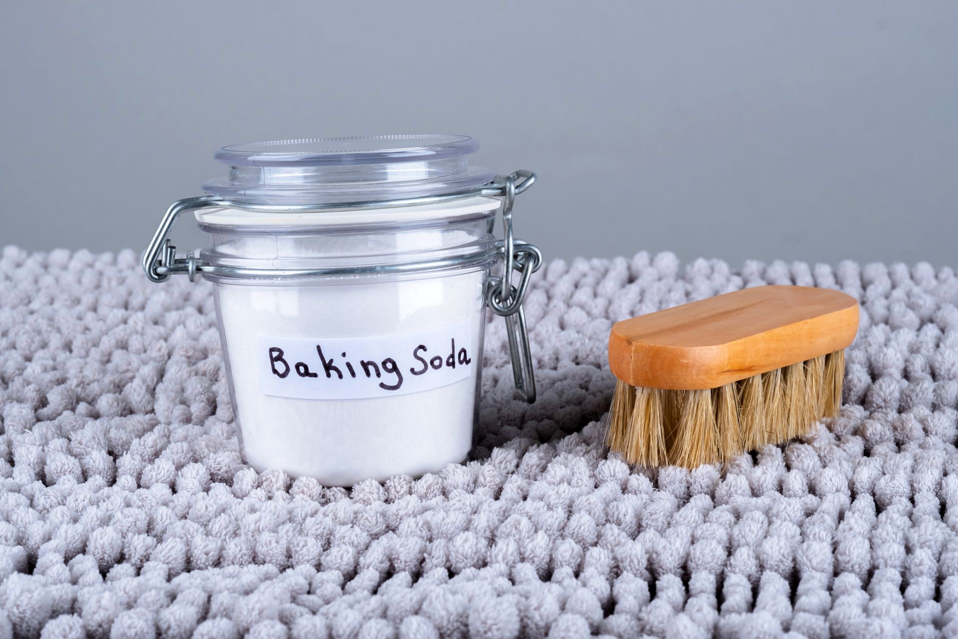 Baking soda and wooden cleaning brush placed on a gray home carpet, ready for eco-friendly cleaning.
