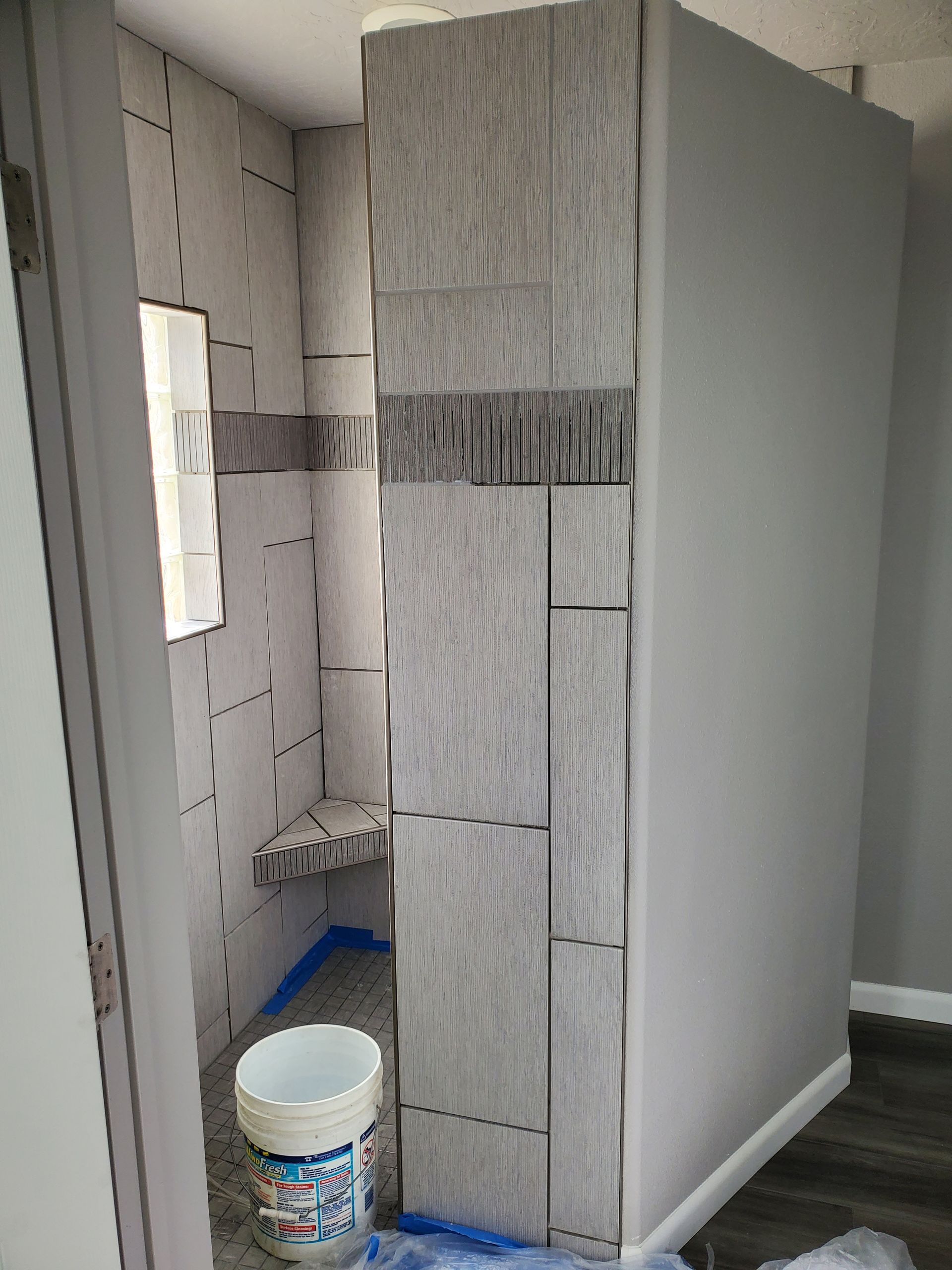 Bathroom with gray and beige tiled shower and wall with a bucket on the floor.