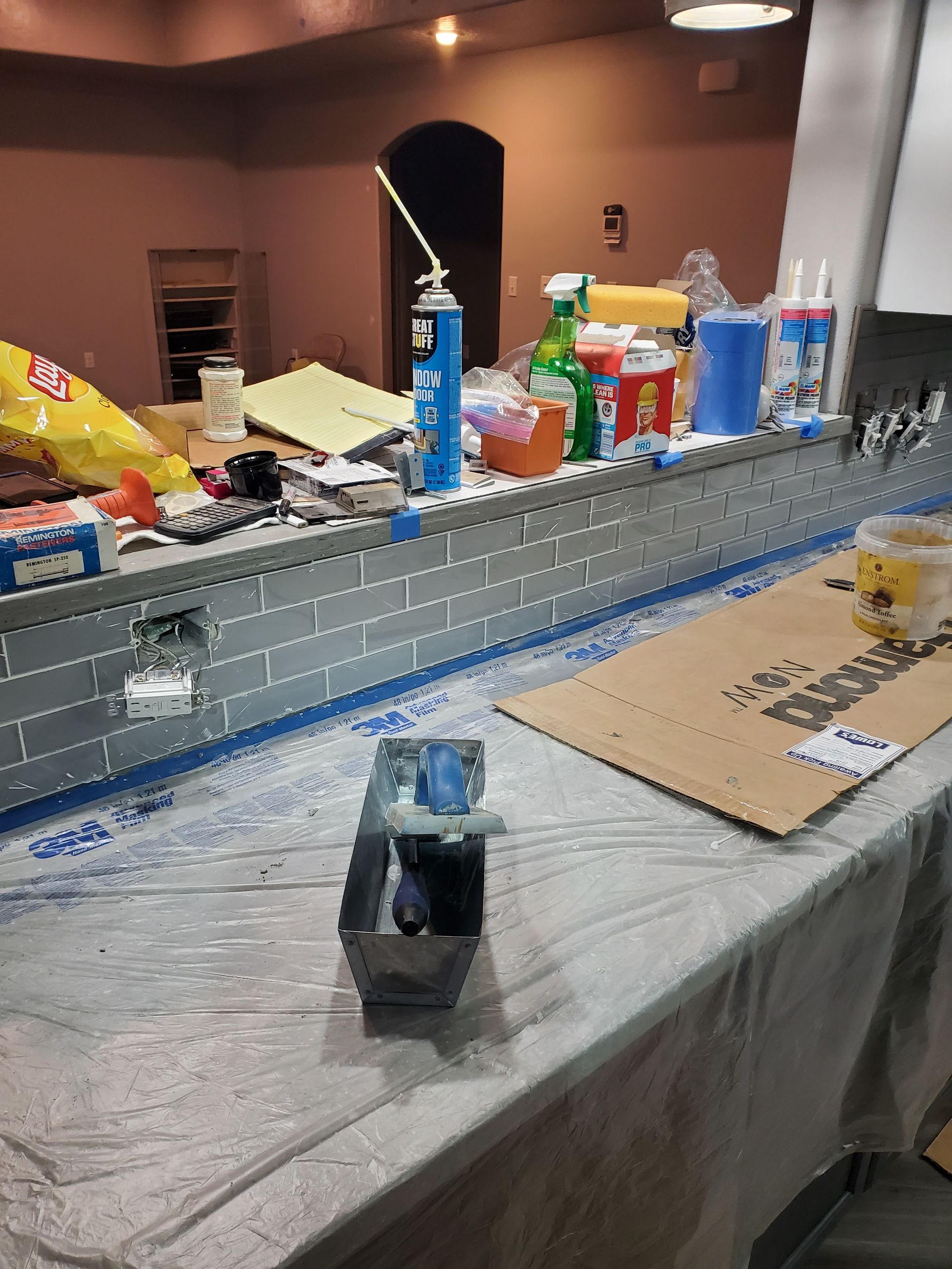 A long, gray brick structure being worked on indoors, with tools and materials on a table covered in plastic.