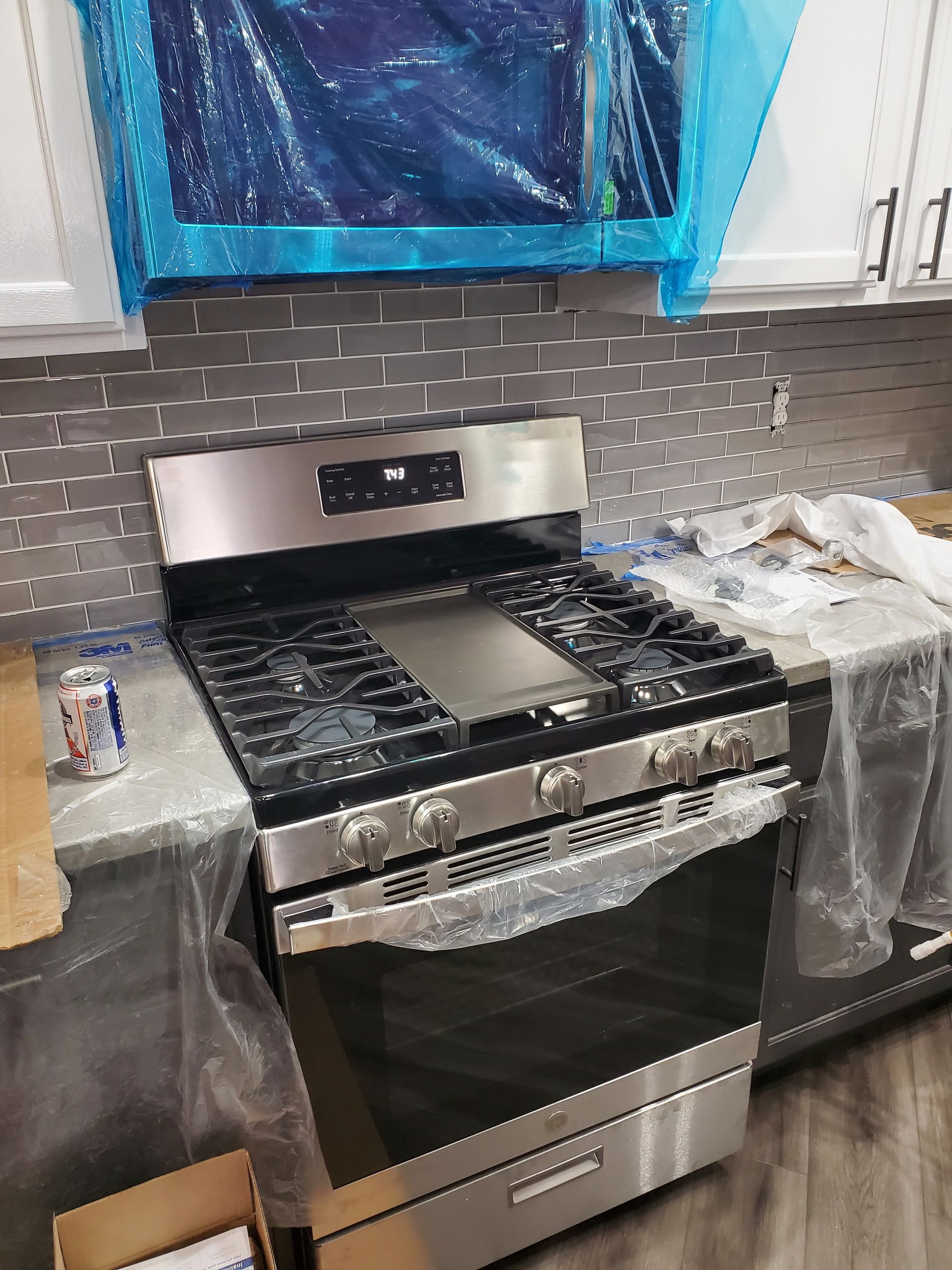 Stainless steel gas range in new kitchen, protective film on oven. Grey countertop, tiled backsplash.
