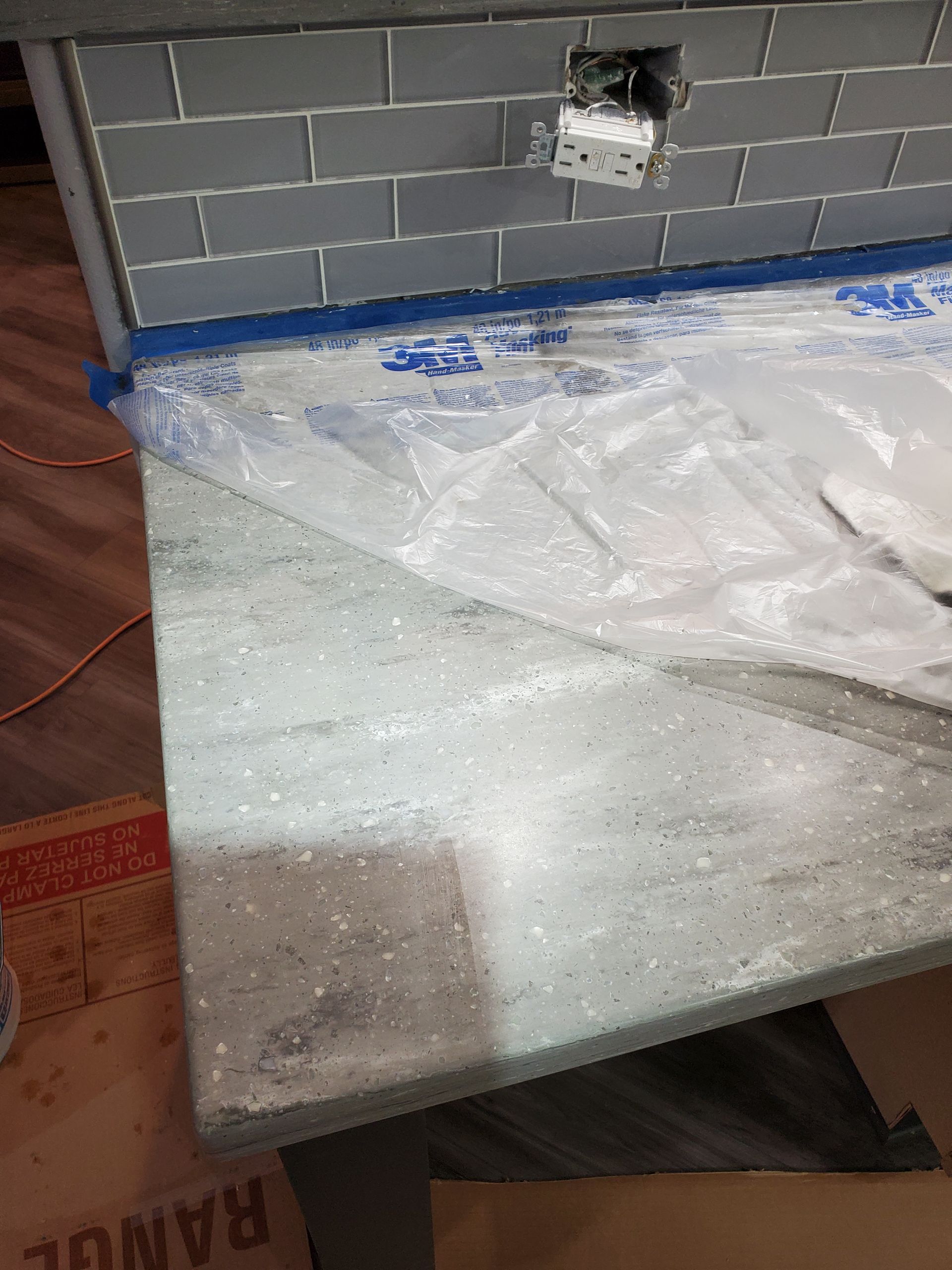 A countertop with protective plastic, against gray tiled backsplash. An electrical outlet is visible.