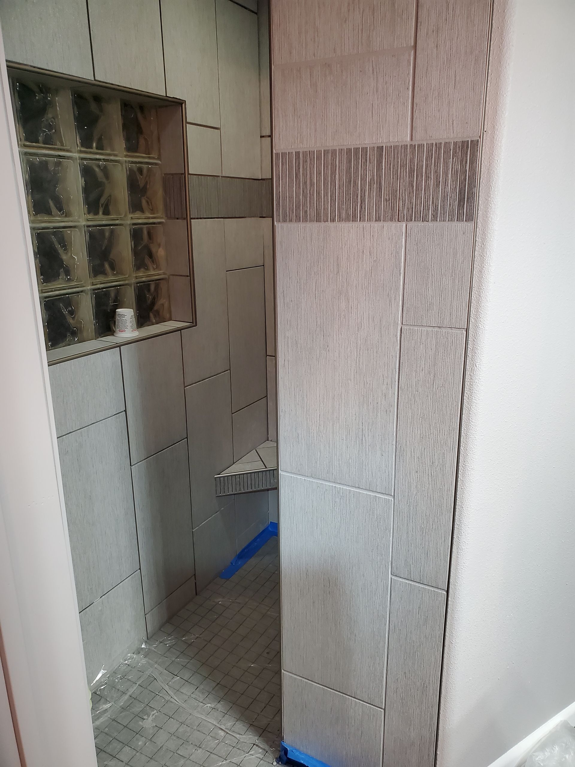 Bathroom under renovation with gray tiled walls, a glass block window, and exposed plumbing.