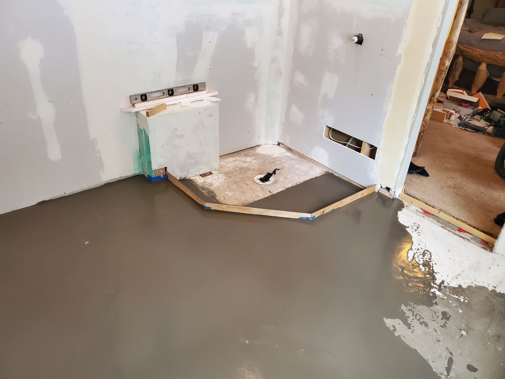 Room under construction with wet, grey concrete floor. Shower base framed with wood. White walls.