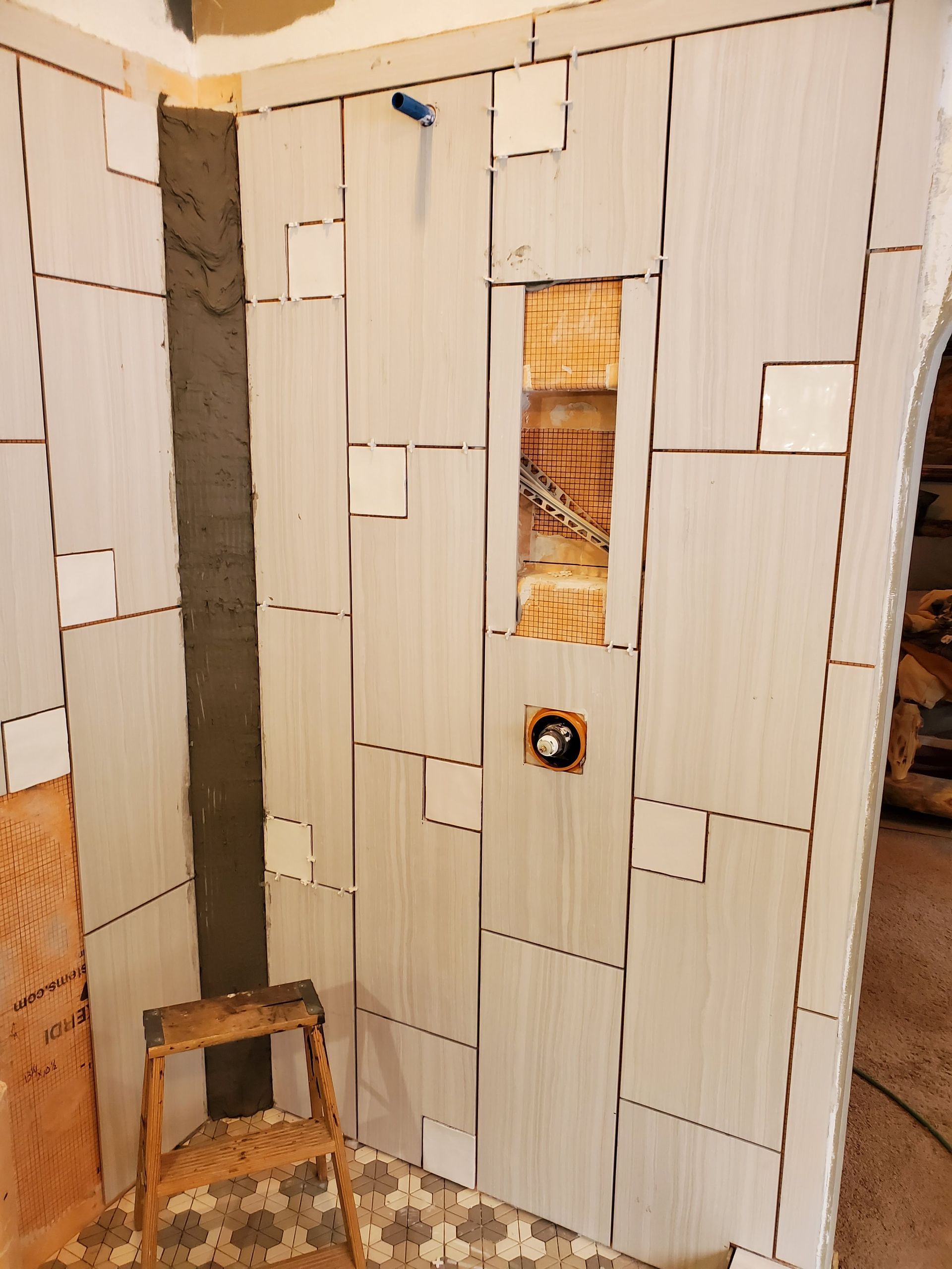Shower wall under construction with large, light-colored tiles and smaller square accents. A recessed shelf is visible.