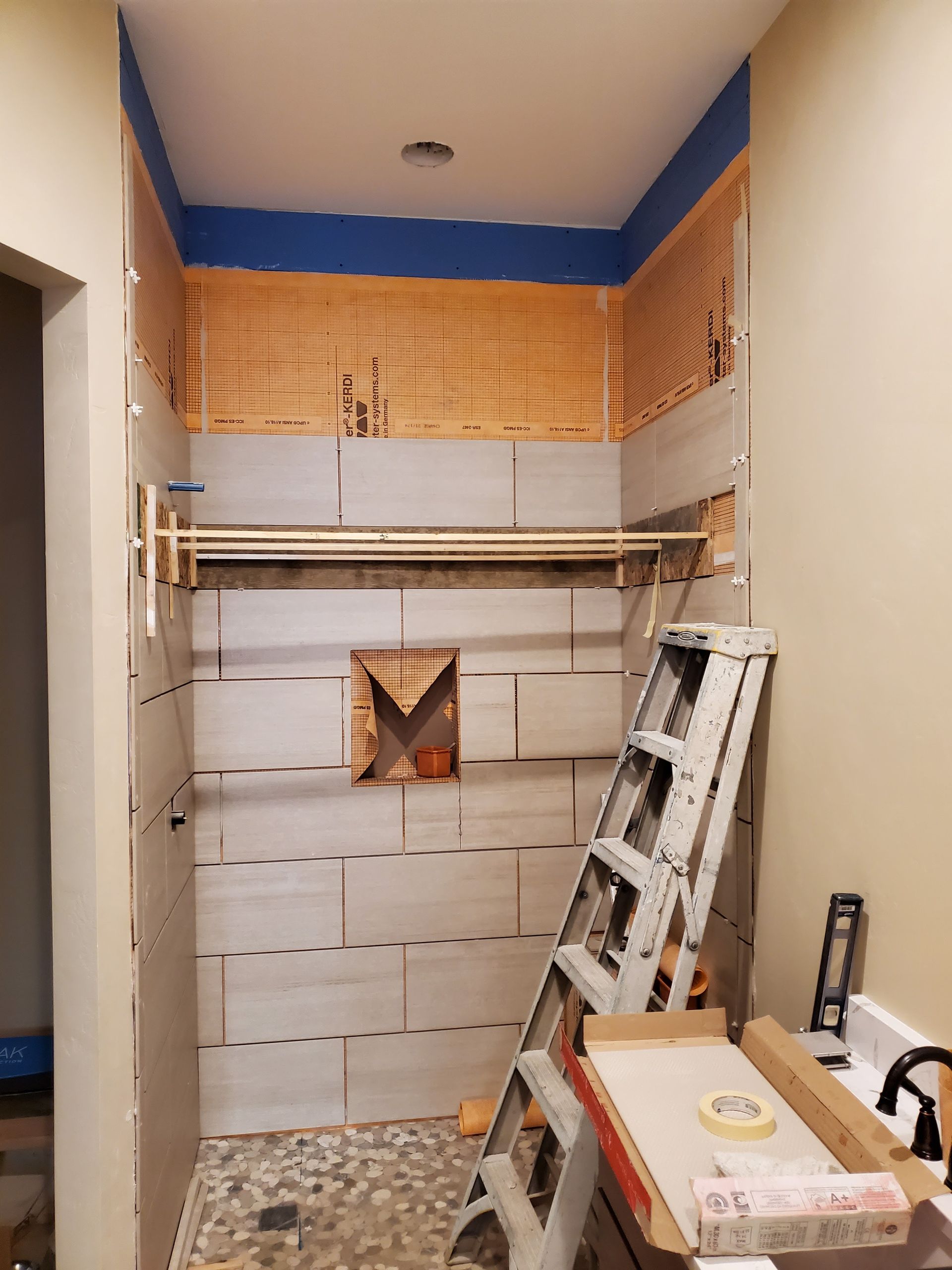 Shower construction in progress, with tiled walls, pebble floor, and ladder inside.