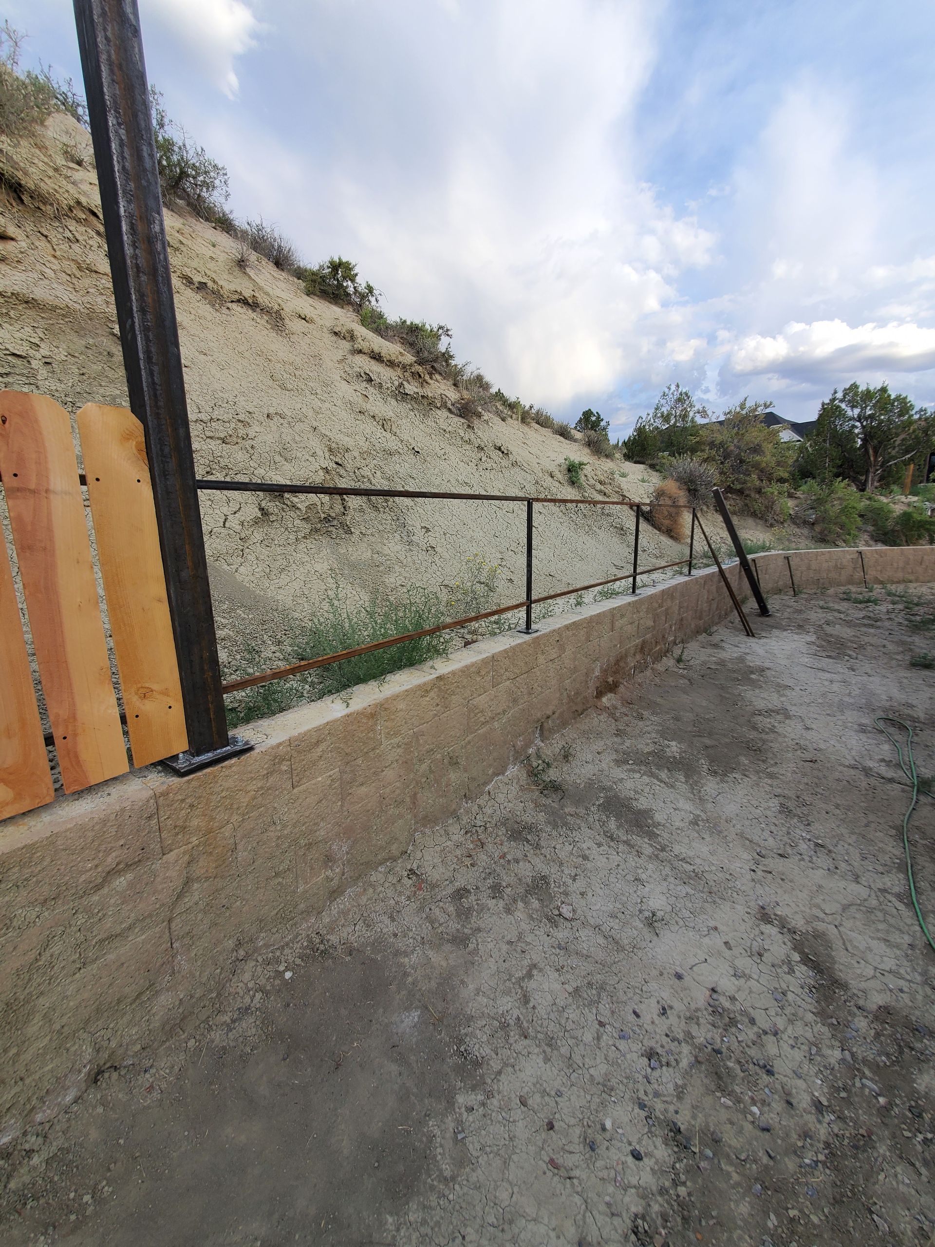 A low concrete retaining wall with a black railing along a hillside.  A wooden fence post is visible.