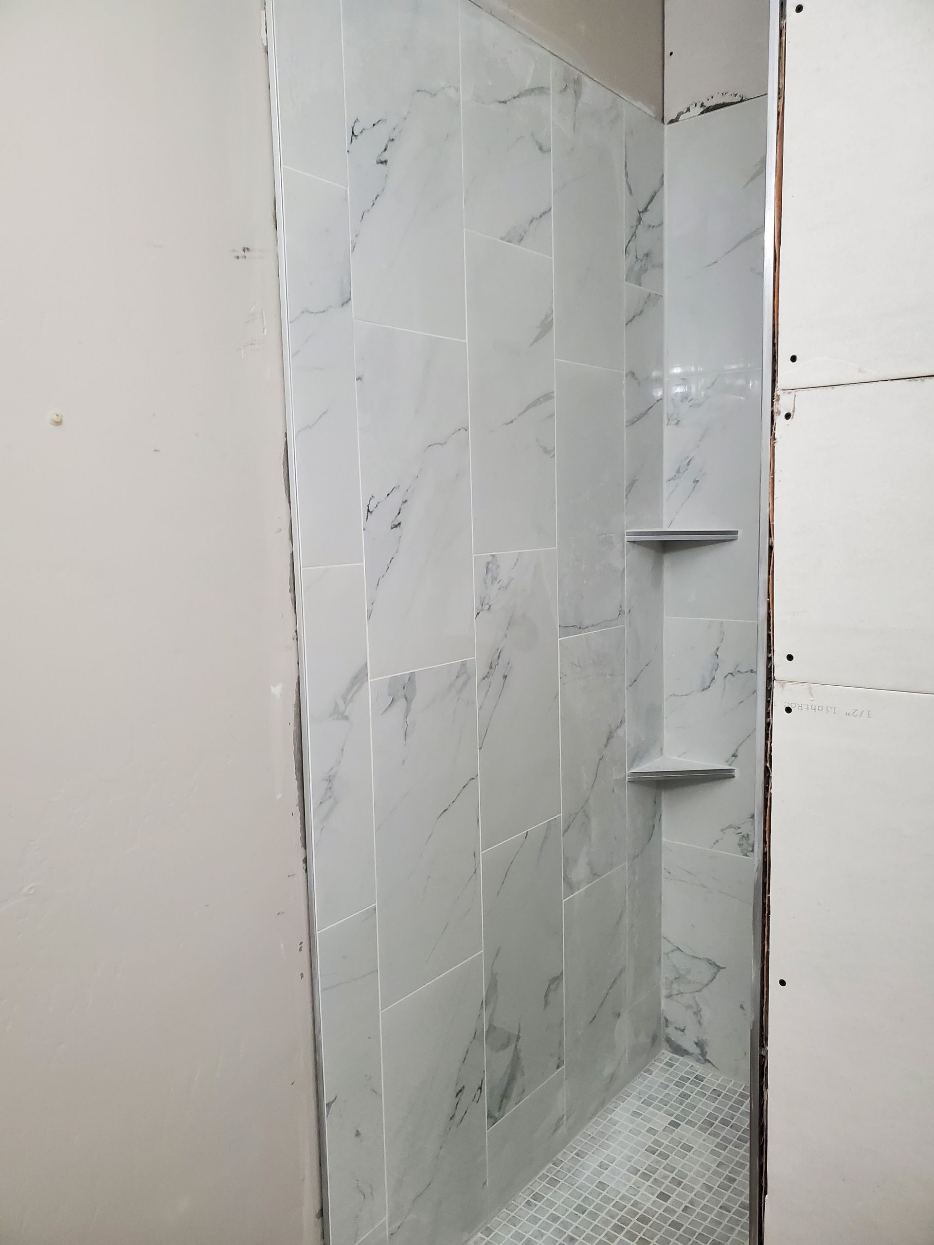 A partially tiled shower stall with two built-in shelves. Walls are white with gray veining.