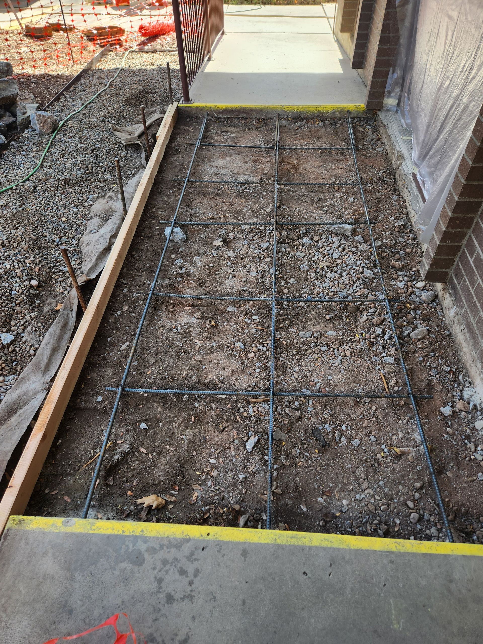 Construction site: rectangular area with rebar grid prepared for concrete walkway. Wooden forms and exposed dirt are visible.