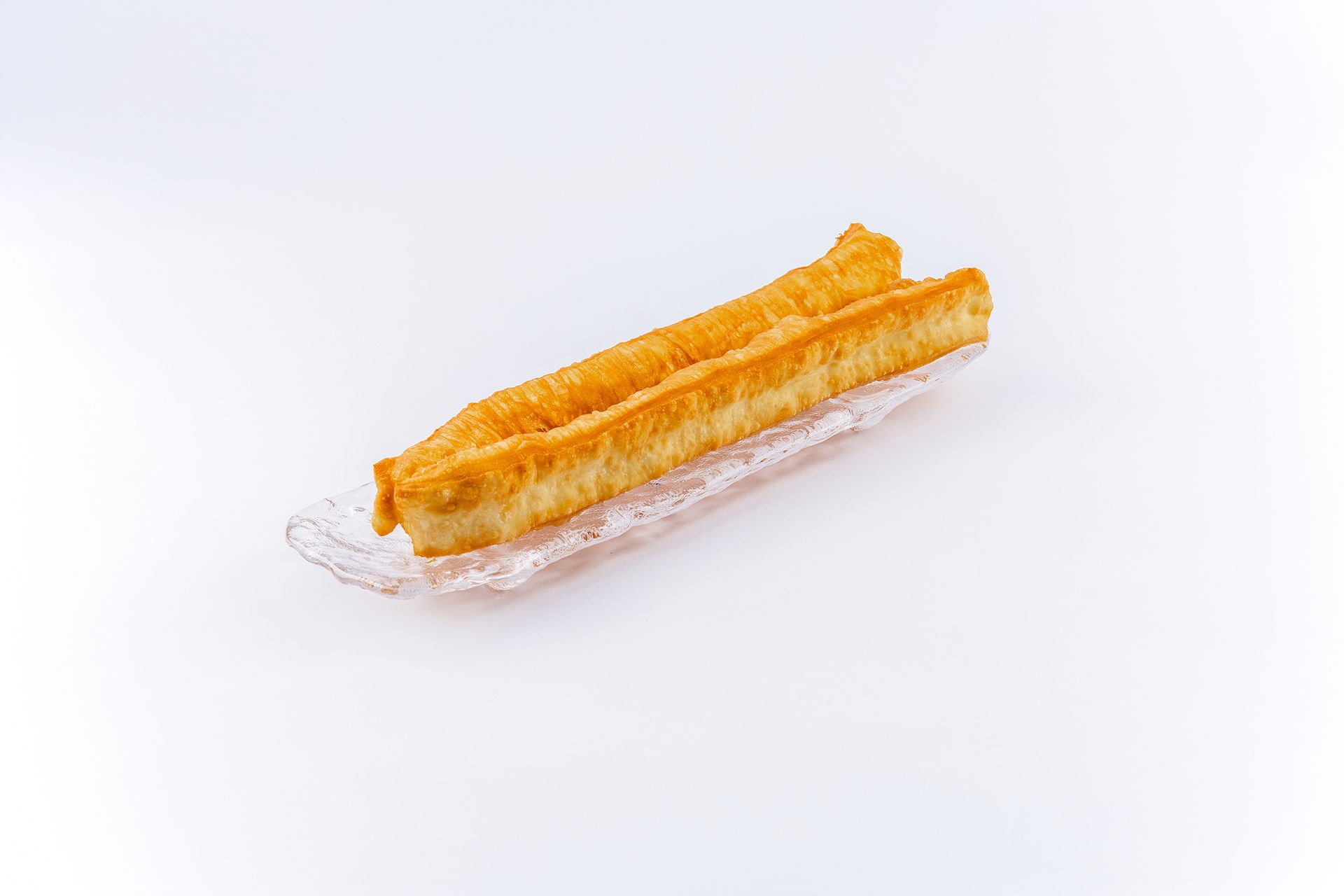 A long, golden-brown deep-fried dough stick (youtiao) sitting on a piece of clear plastic wrap against a white background.